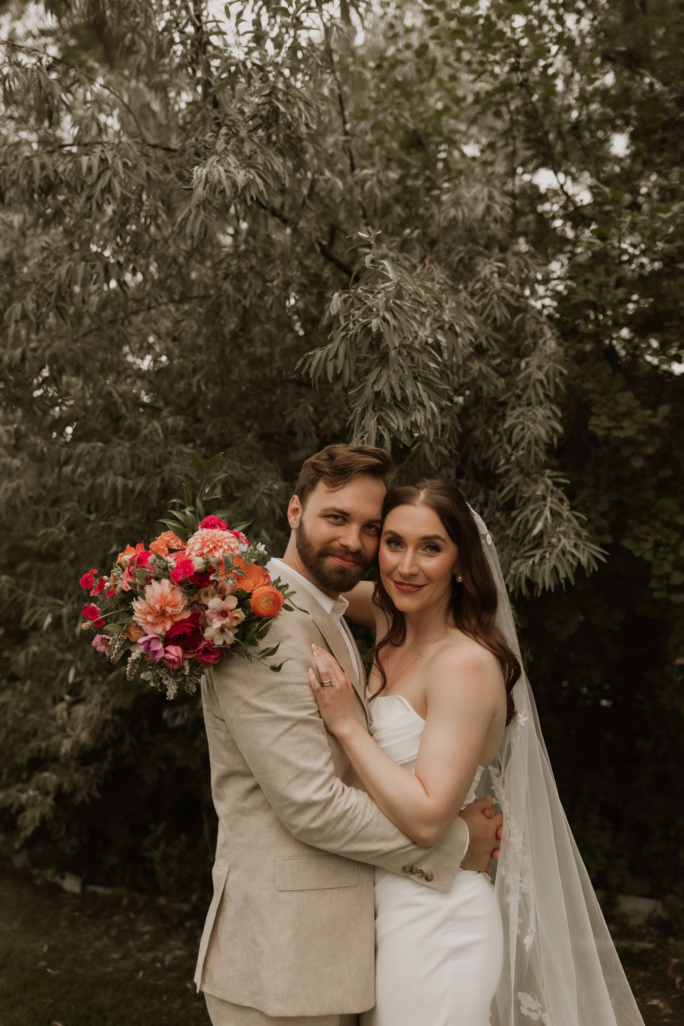 Romantic garden wedding portrait with couple holding vibrant pink and orange floral bouquet.