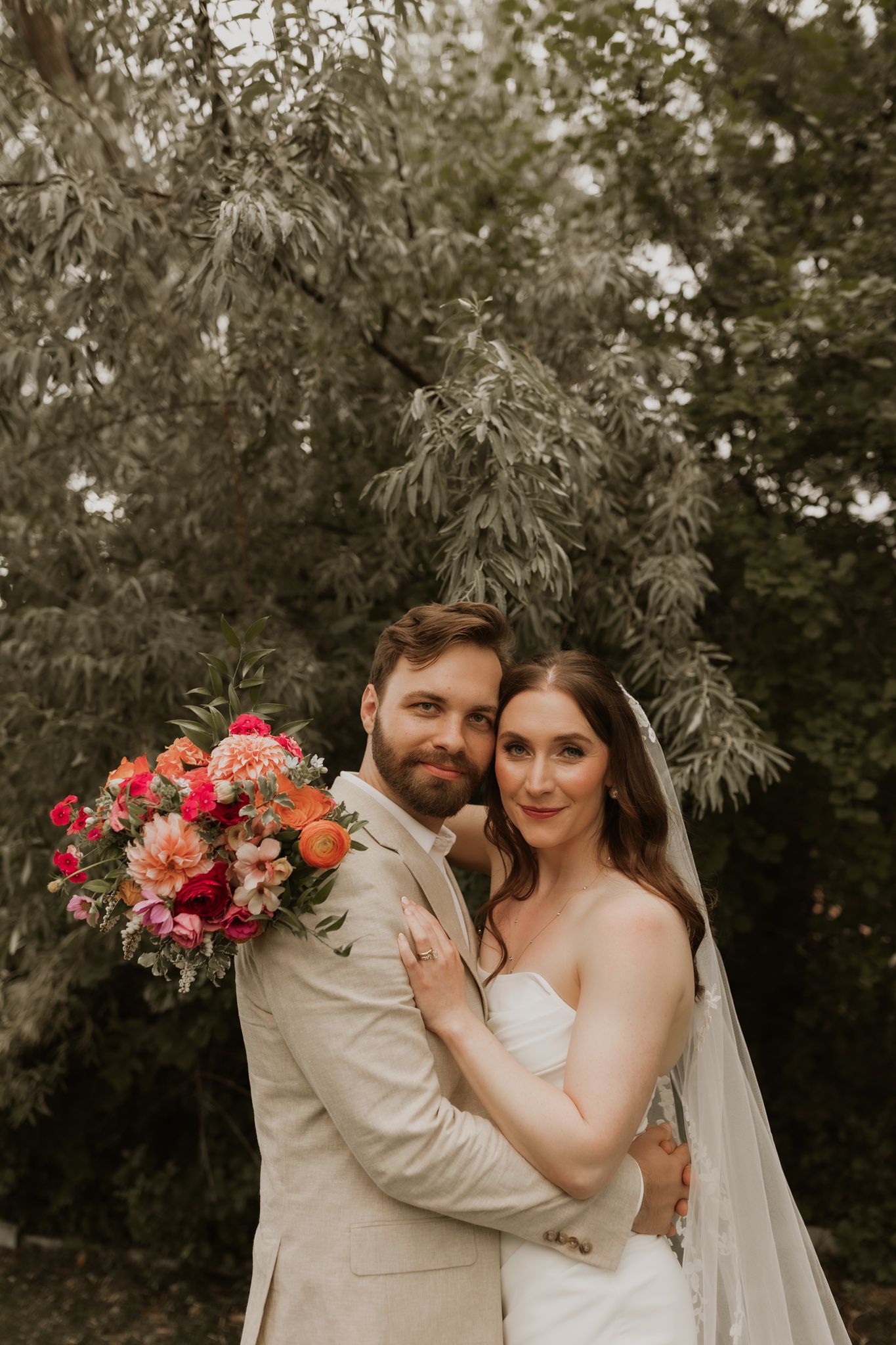 A romantic wedding photo of a couple embracing outdoors with vibrant coral and pink floral bouquet.