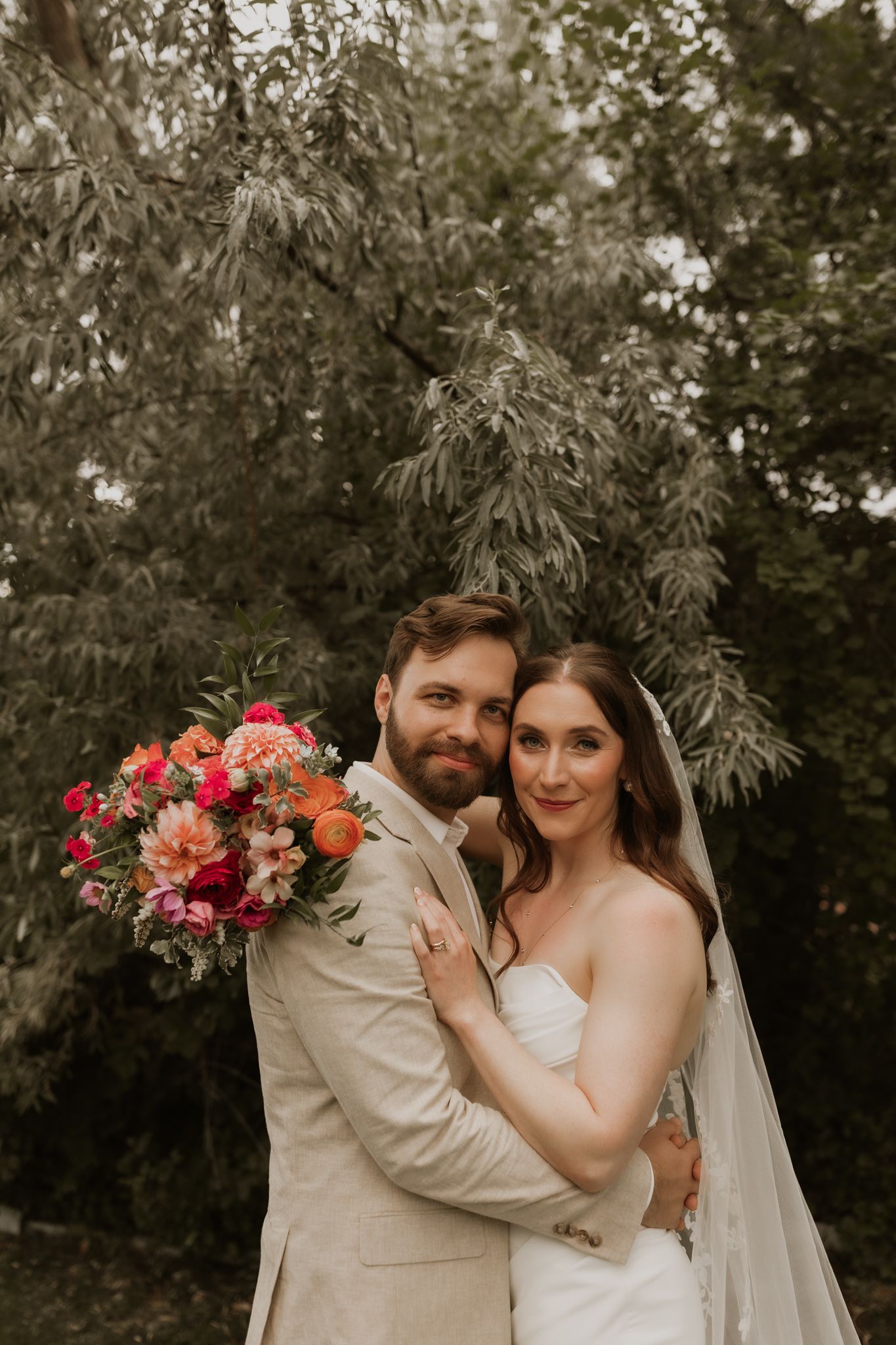 Wedding couple poses with colorful bouquet against backdrop of lush trees.