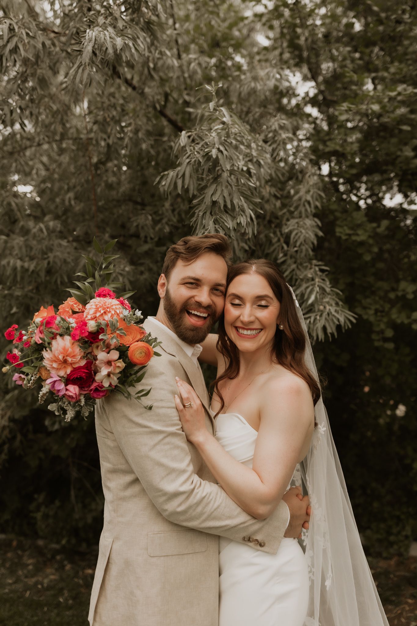 A joyful couple embraces while holding a vibrant coral and pink bouquet against a lush garden backdrop.