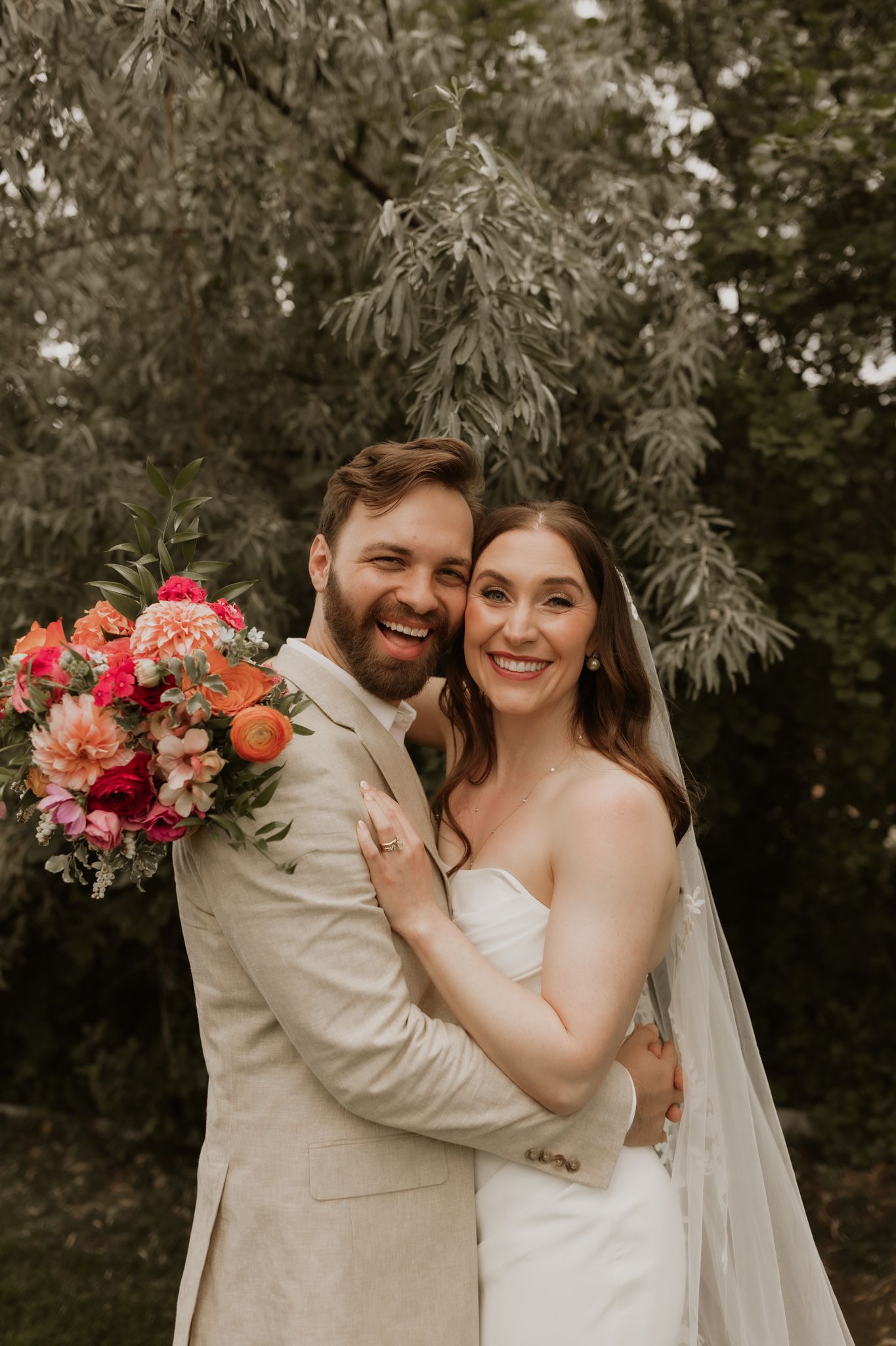 Joyful wedding couple sharing a laugh while holding a bright coral and pink bouquet.