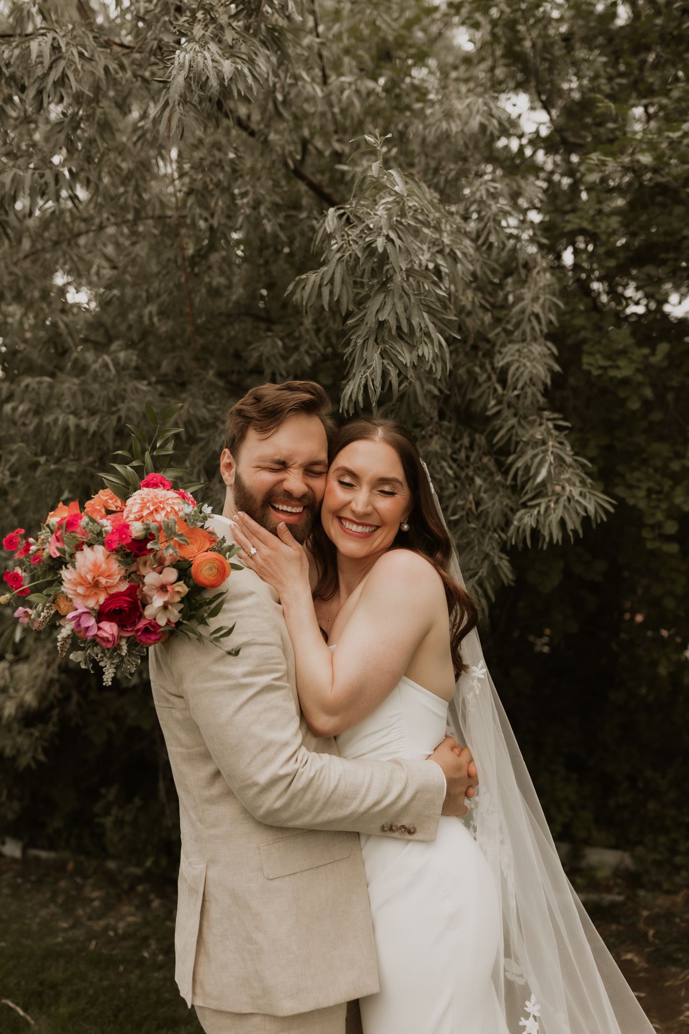 A romantic wedding photo showing a couple embracing outdoors with a vibrant coral and pink bouquet.