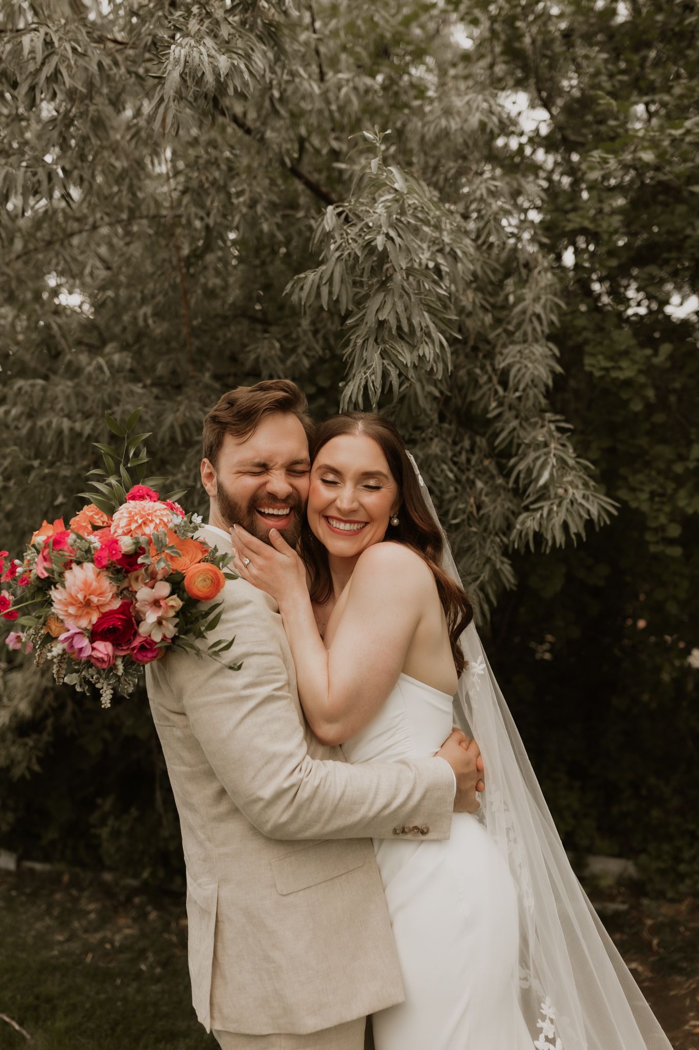A romantic wedding photo showing a couple embracing outdoors with vibrant coral and pink florals against a natural backdrop.