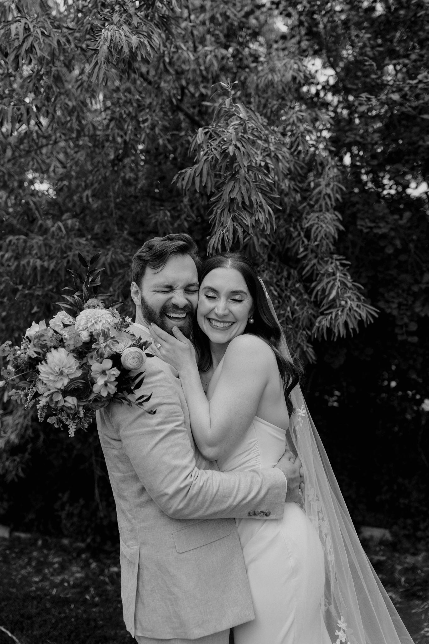 A romantic black and white wedding photo of a couple embracing outdoors with trees in the background.