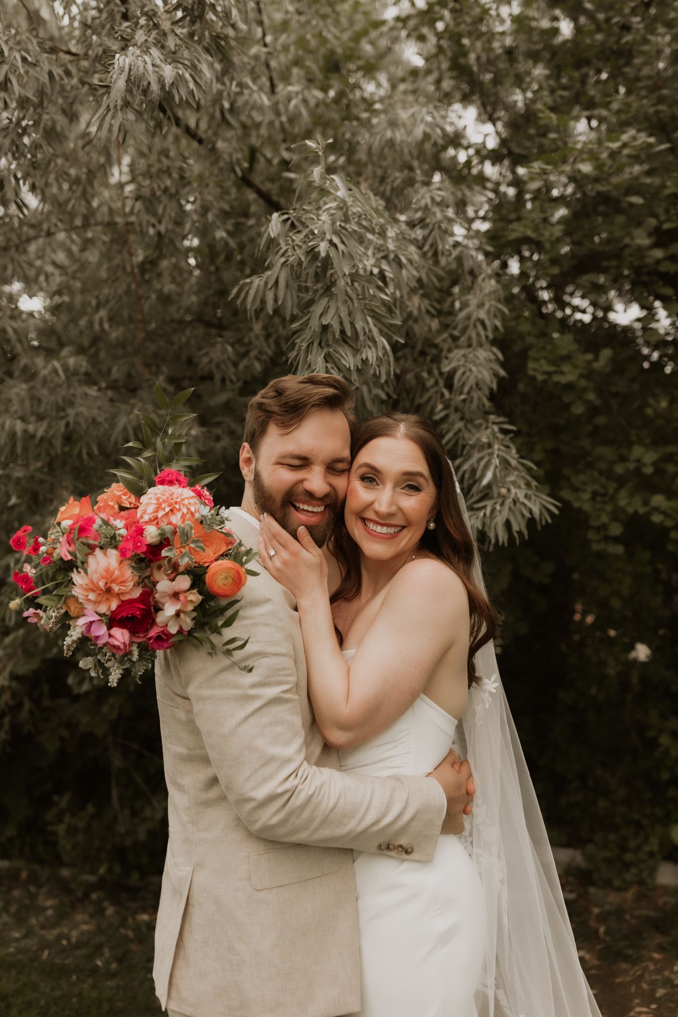 A joyful wedding couple embraces outdoors surrounded by greenery with a vibrant coral and pink bouquet.