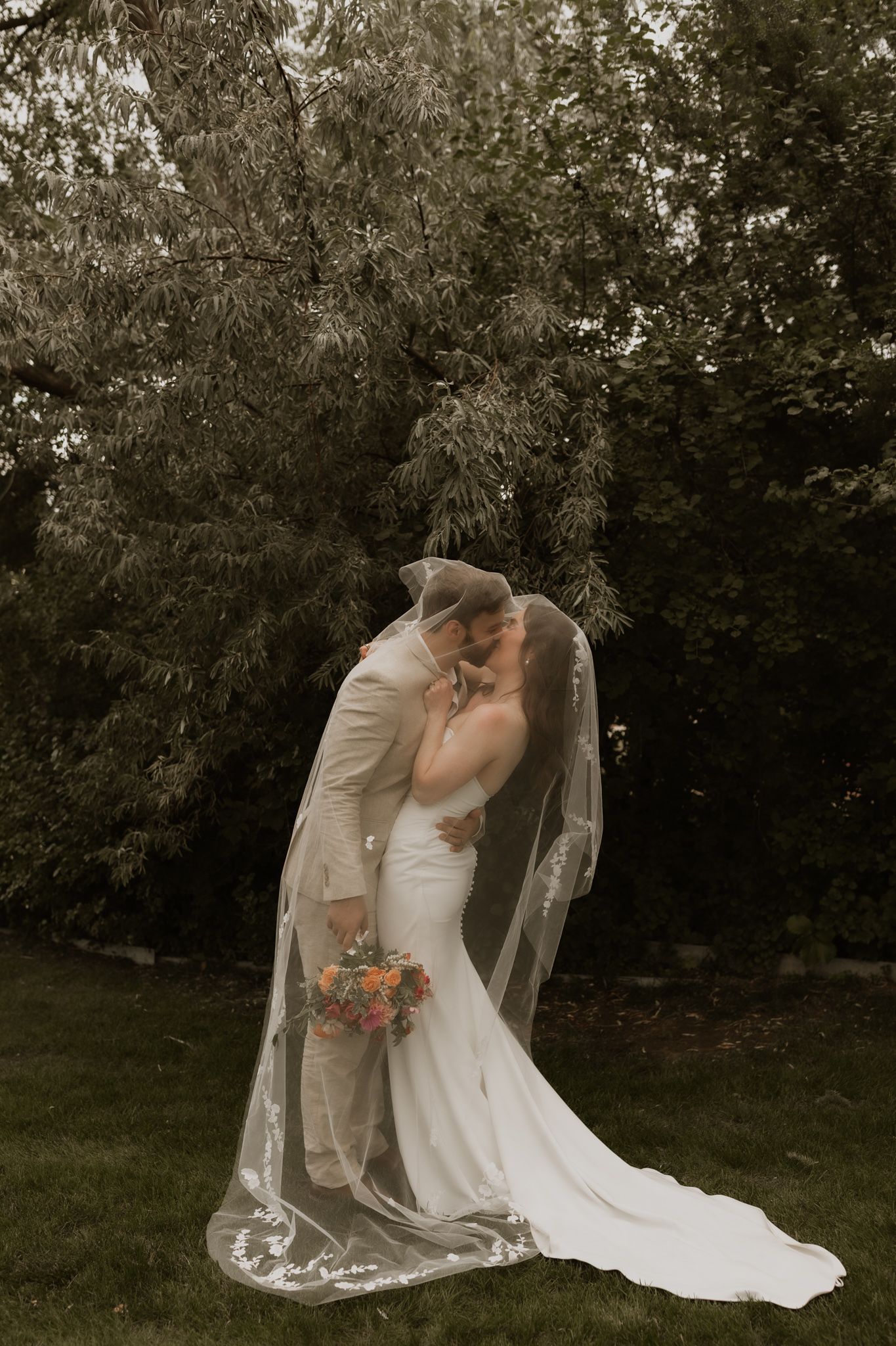 Wedding couple shares intimate moment under trees with flowing white veil and beige suit.