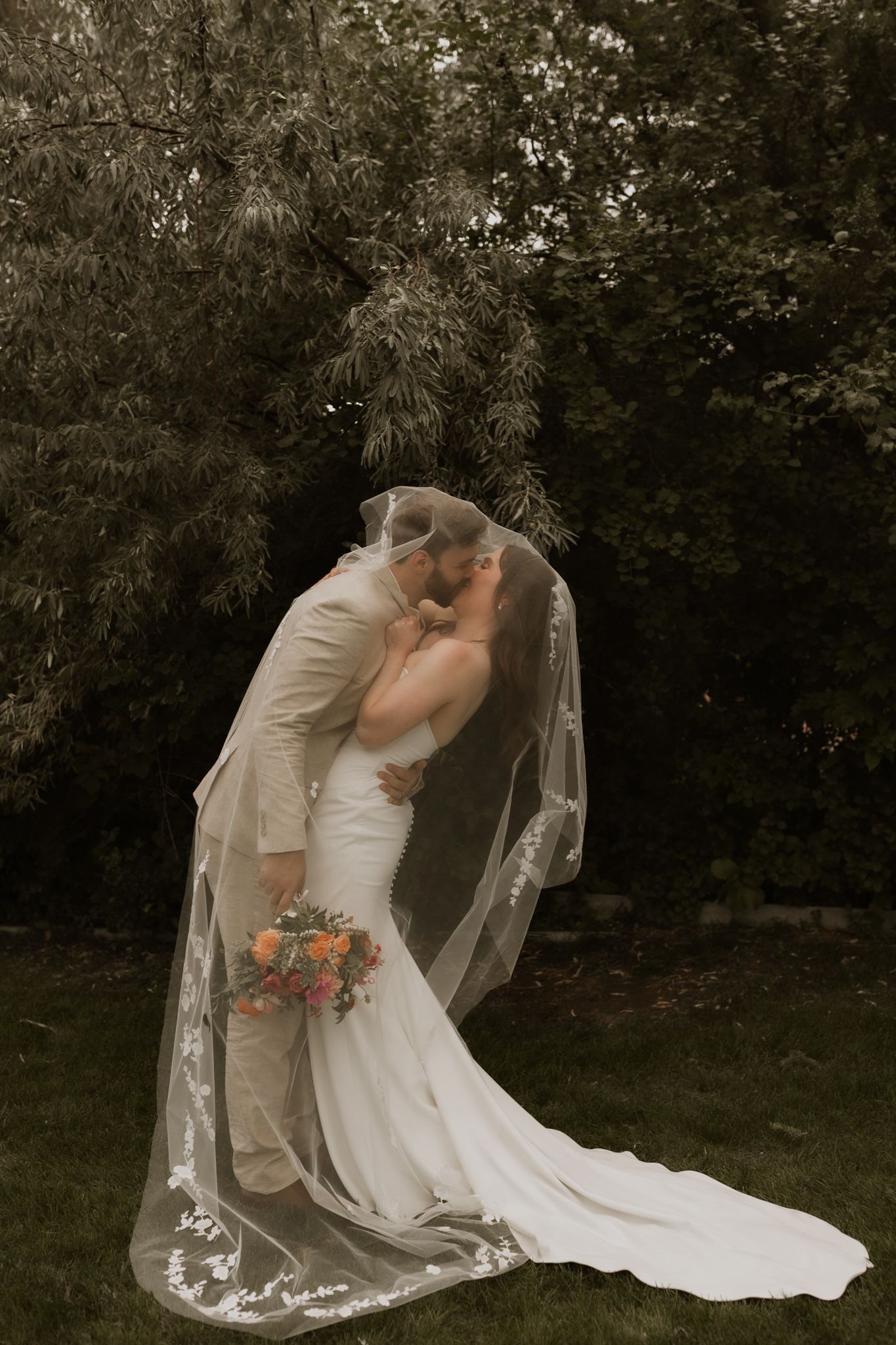 A couple shares an intimate moment under a long flowing wedding veil in an outdoor garden setting.