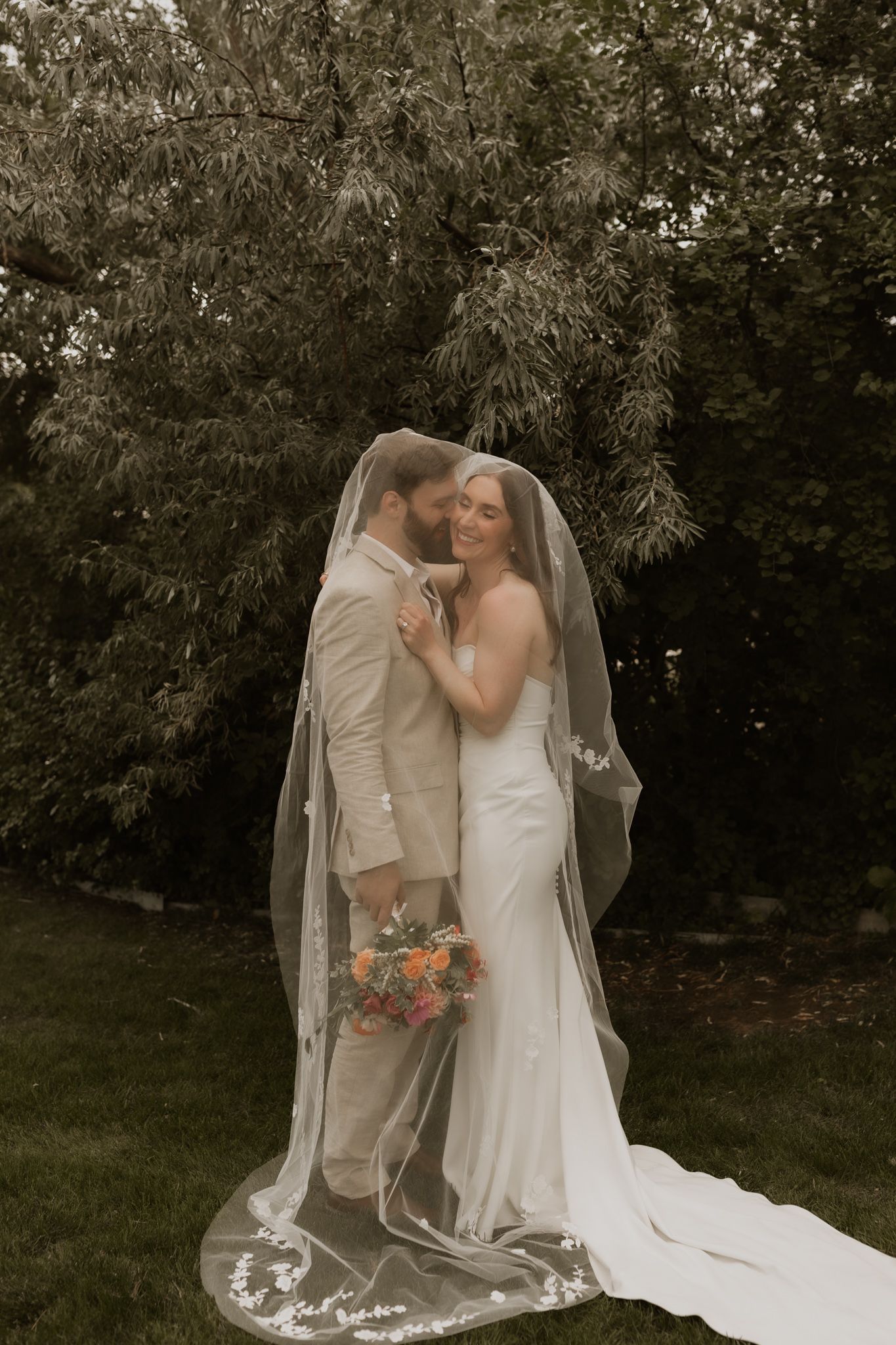 An intimate wedding portrait showing a tender embrace against a natural outdoor backdrop with flowing white veil.