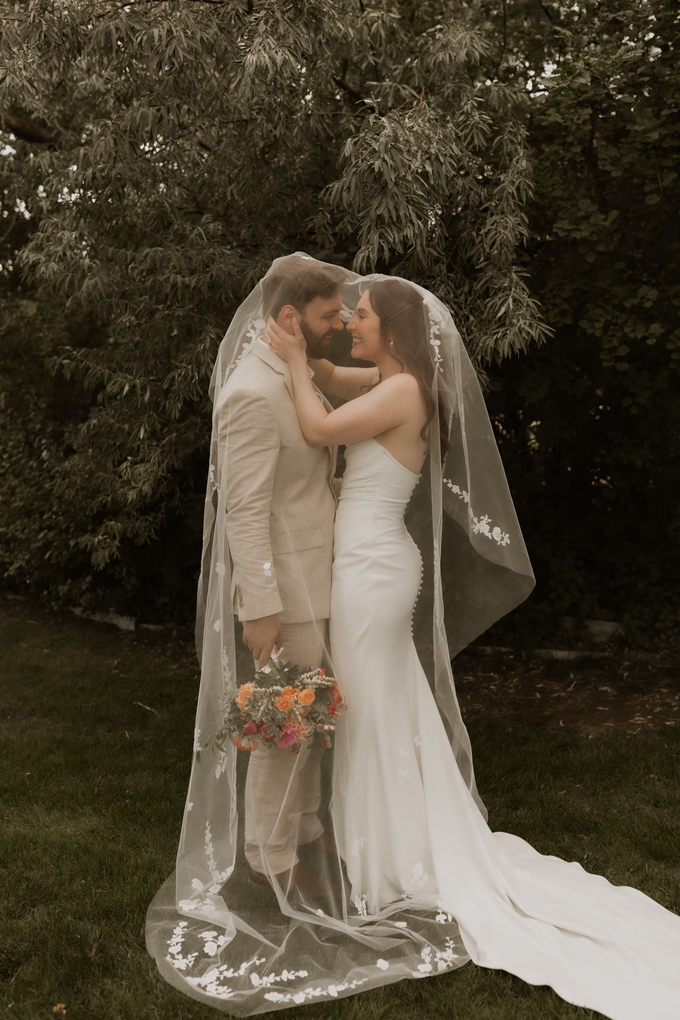 Newlyweds share a tender moment outdoors with the bride's veil flowing in the breeze.