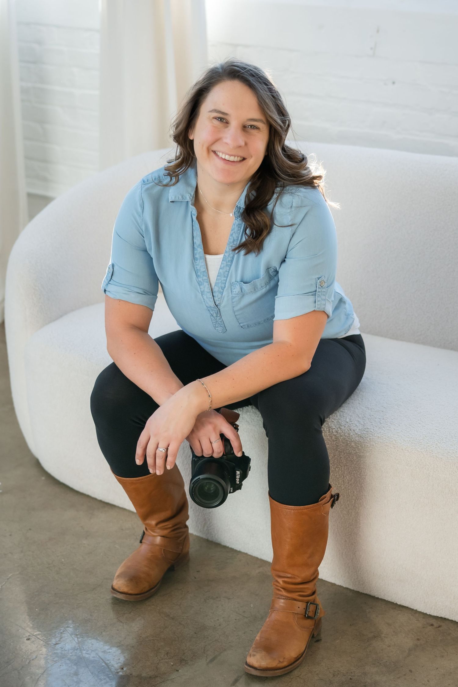 Professional photographer in light blue chambray shirt and brown boots sits casually on white chair holding camera.
