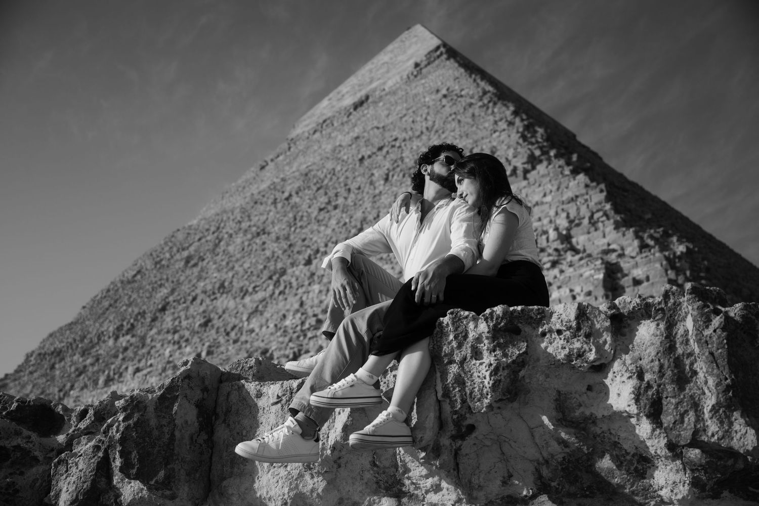 A couple sits together on ancient stone blocks in front of the Great Pyramid of Giza in black and white.