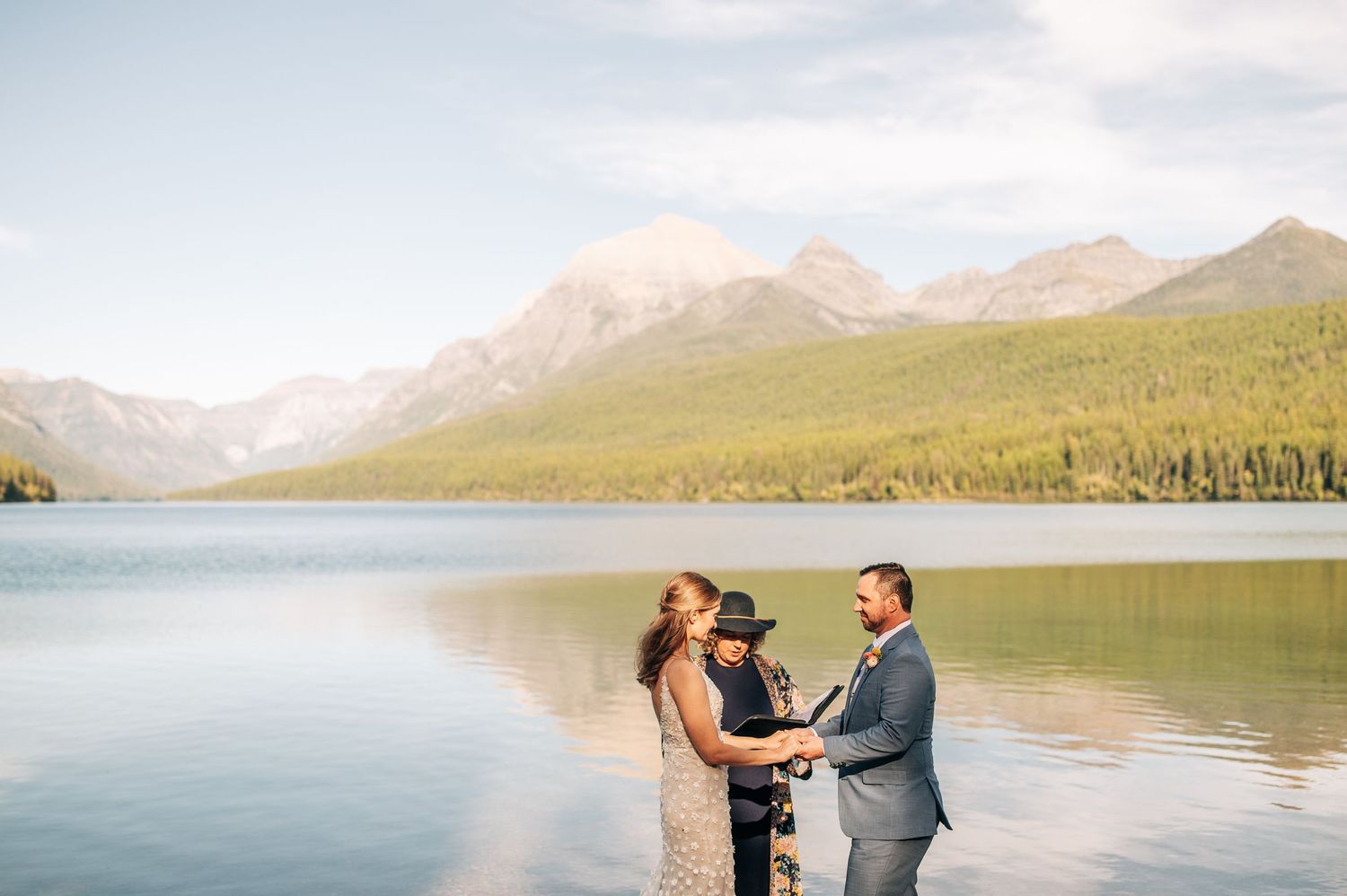 A couple embraces by a serene mountain lake with snow-capped peaks and evergreen forests in the background.