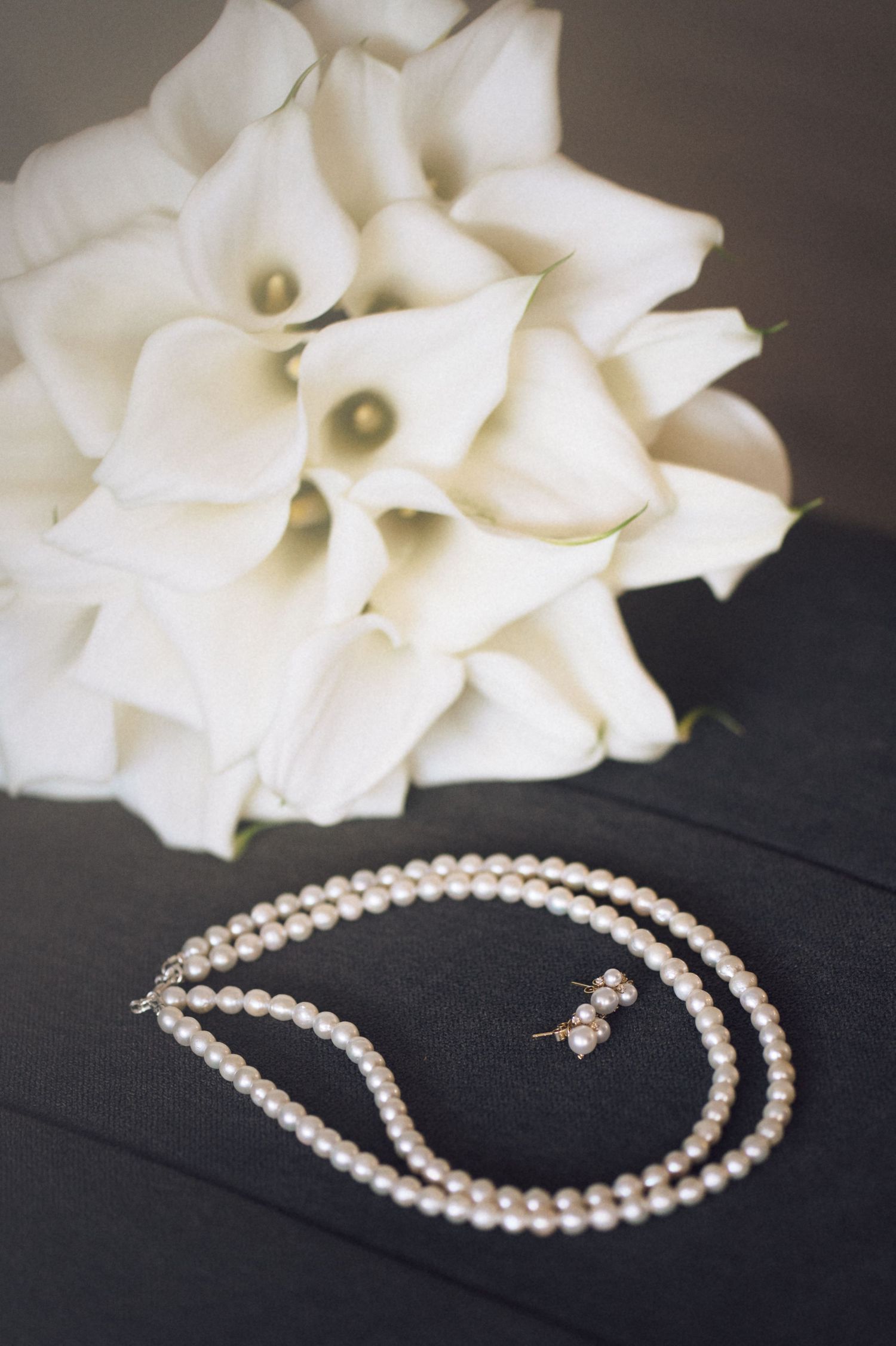 A white calla lily bouquet rests above an elegant pearl necklace and earrings set on a dark surface.