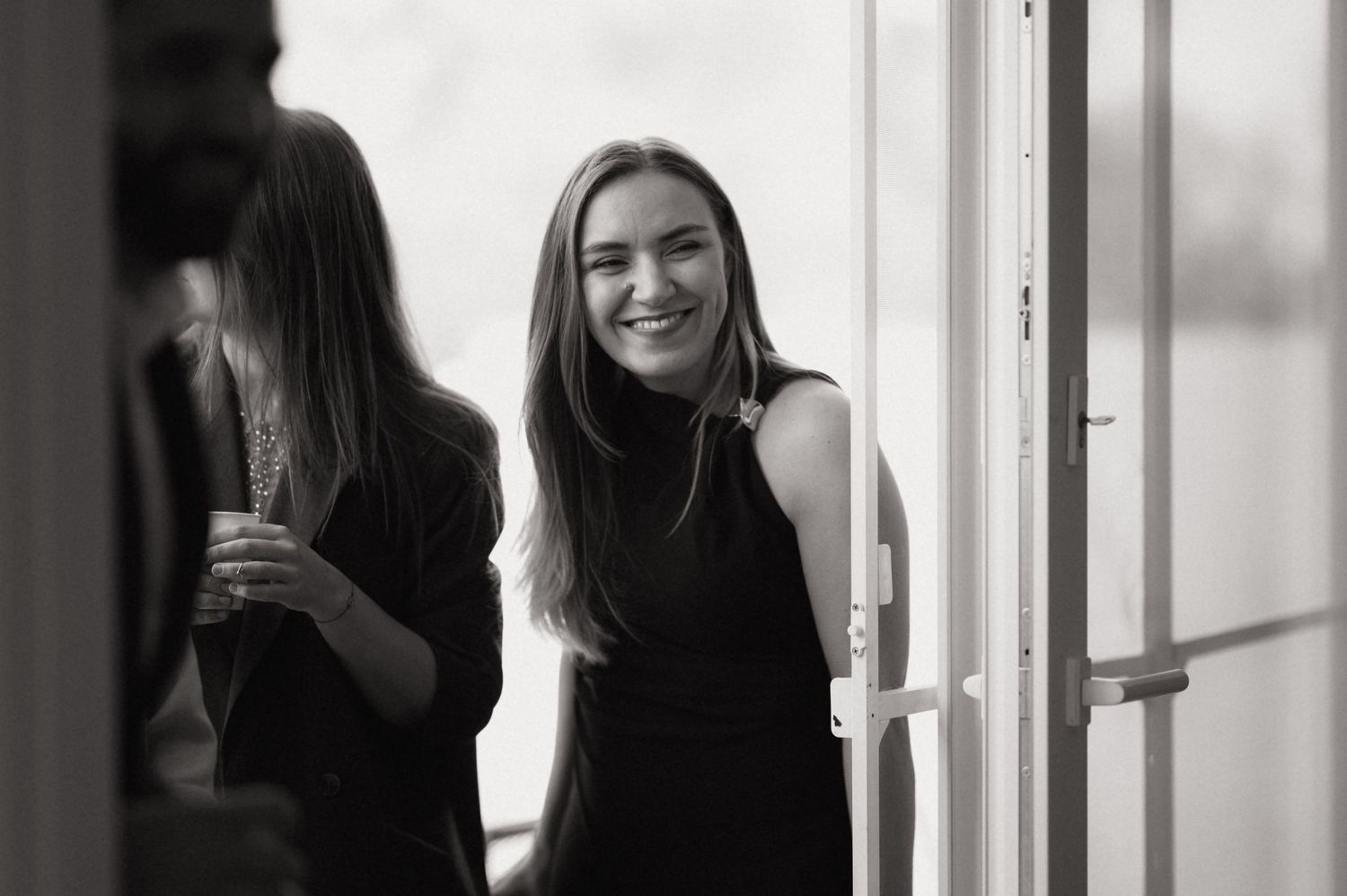 Black and white photo of someone in a sleeveless top smiling by a doorway or window frame.