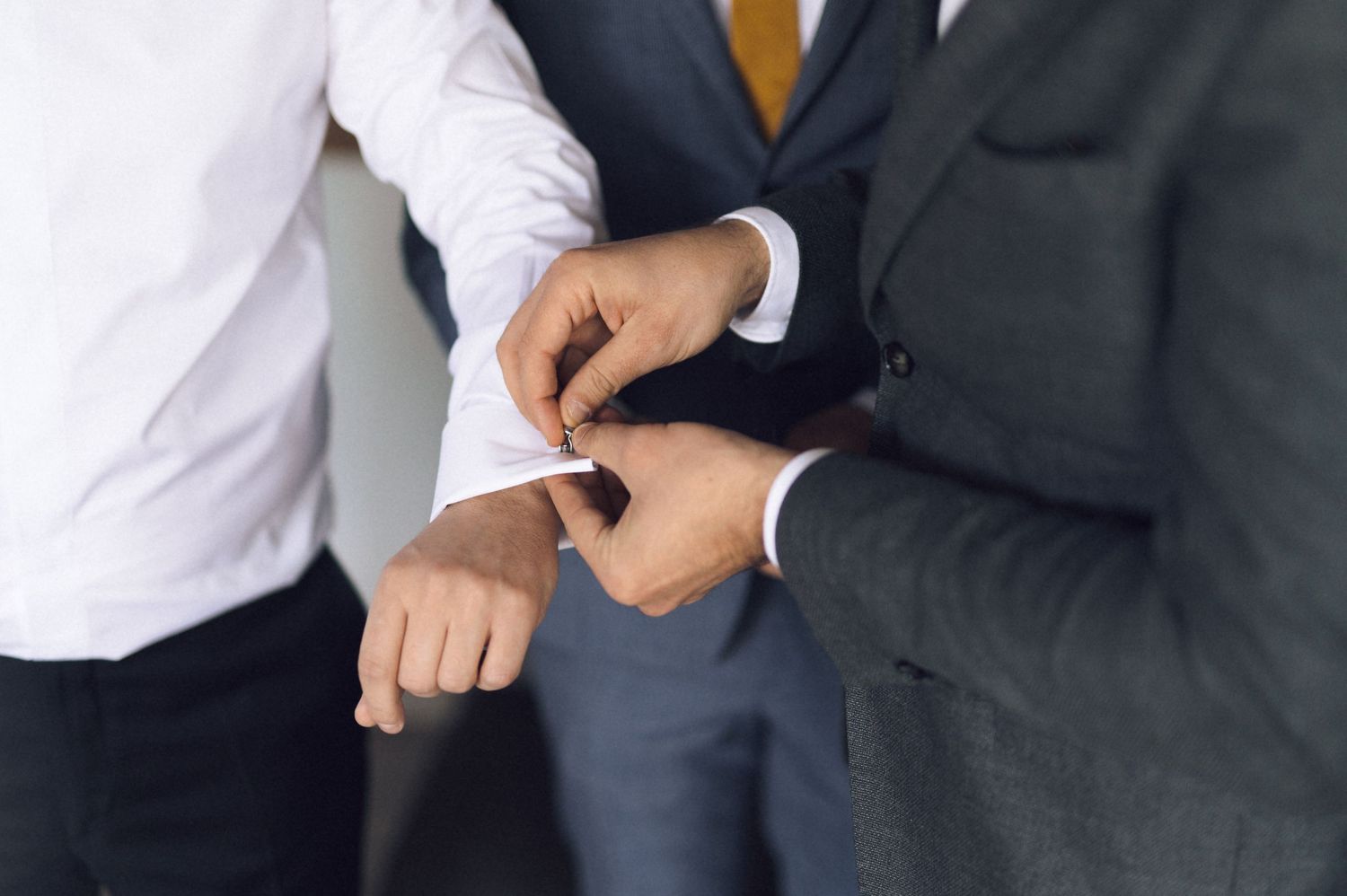 A person in a navy suit and gold tie adjusts their cuff link against a white background.
