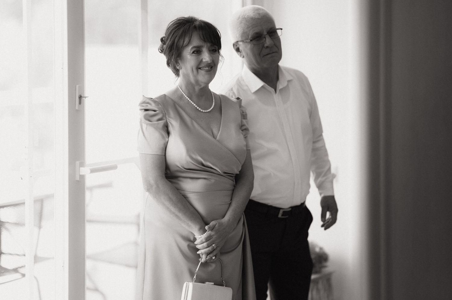 Black and white photo of an elderly couple standing together near a window in casual attire.