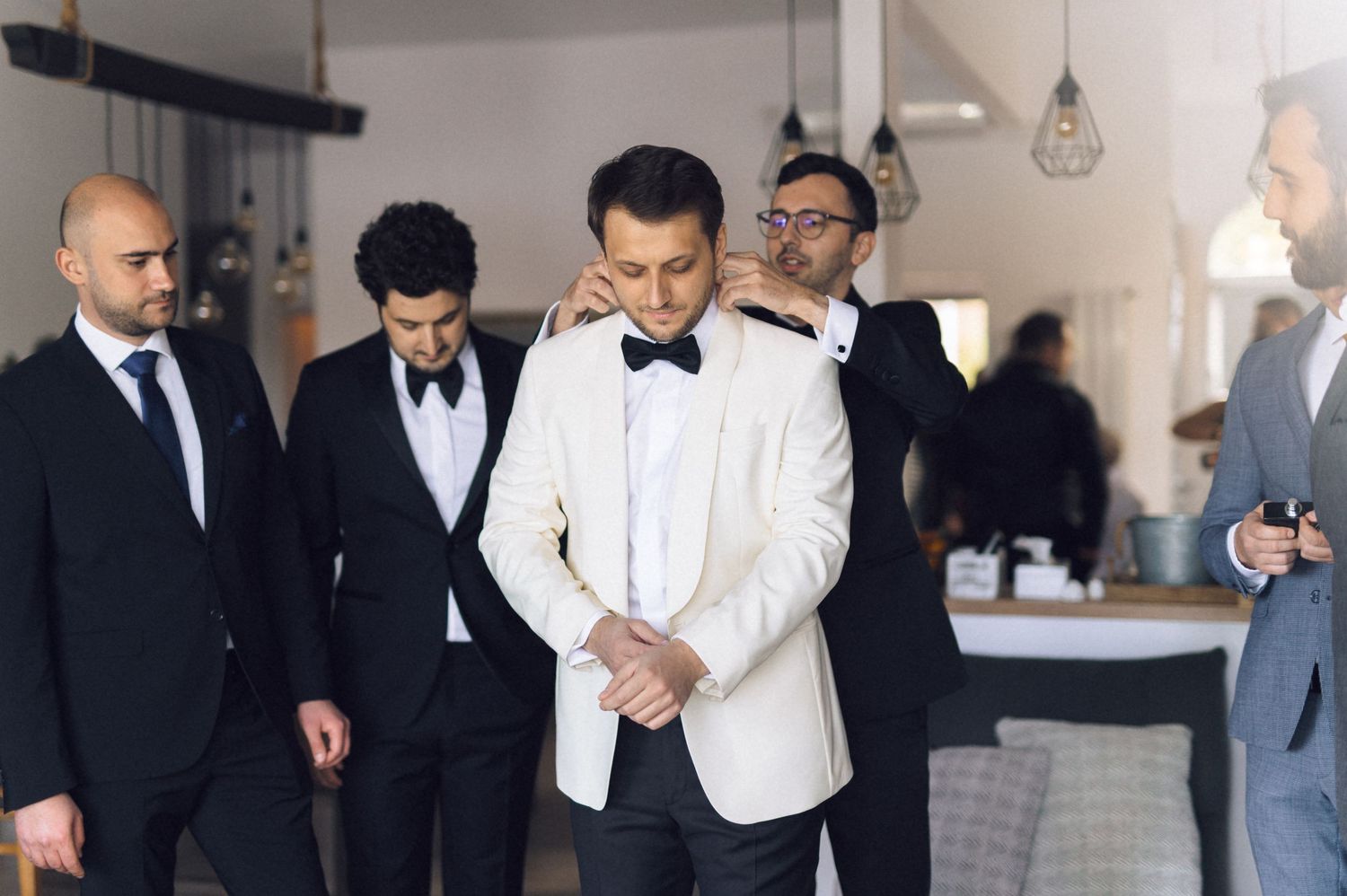 Groomsmen in formal black and white attire getting ready in a bright hotel room before a wedding ceremony.