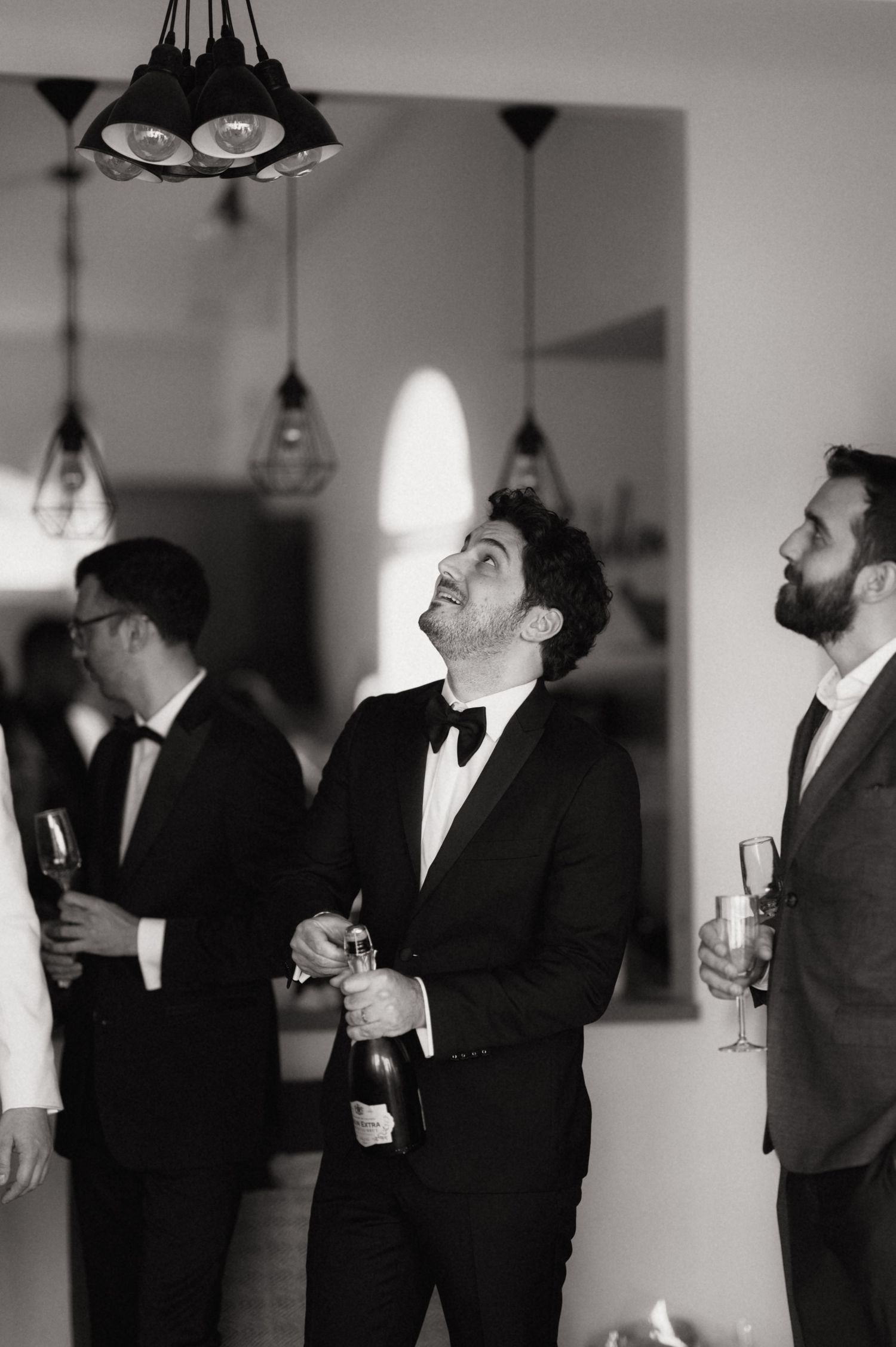 A group of men in formal attire lean back and laugh together at an indoor event in black and white photography.