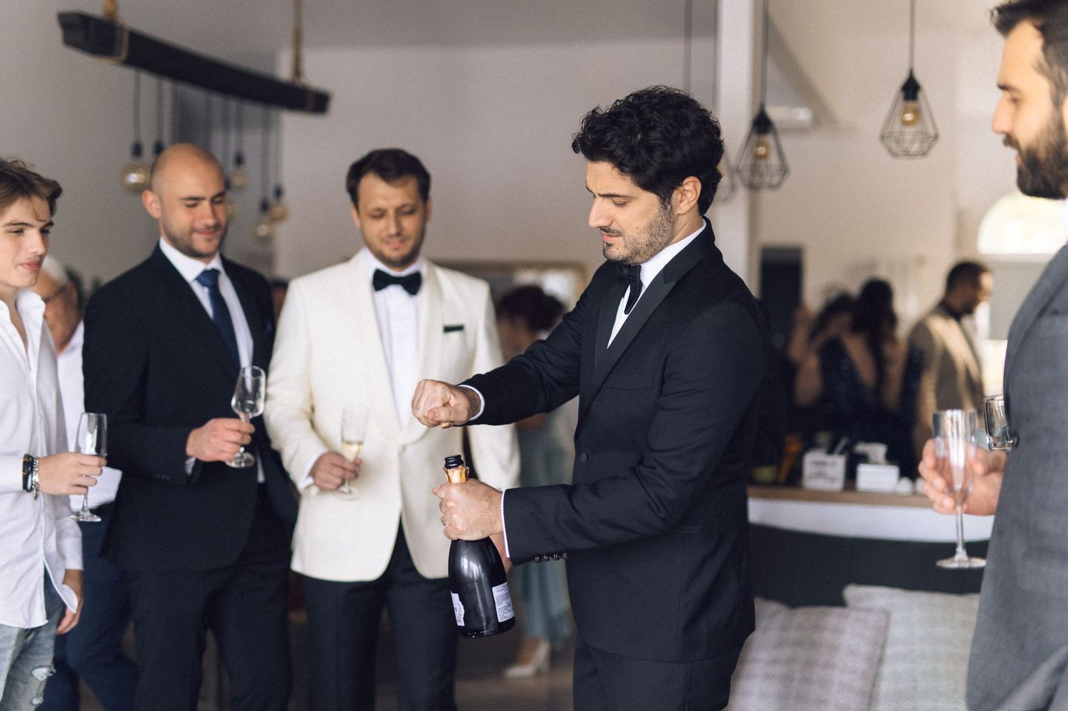A group of well-dressed men in suits and tuxedos gather for drinks at an elegant event with pendant lighting.