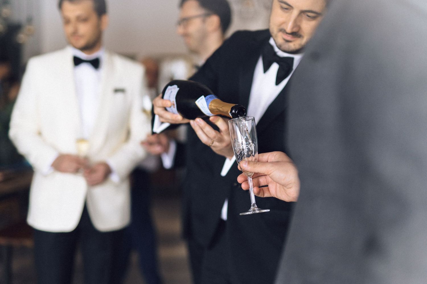 A person in a black tuxedo pours champagne into a glass at an elegant event with others in formal attire.
