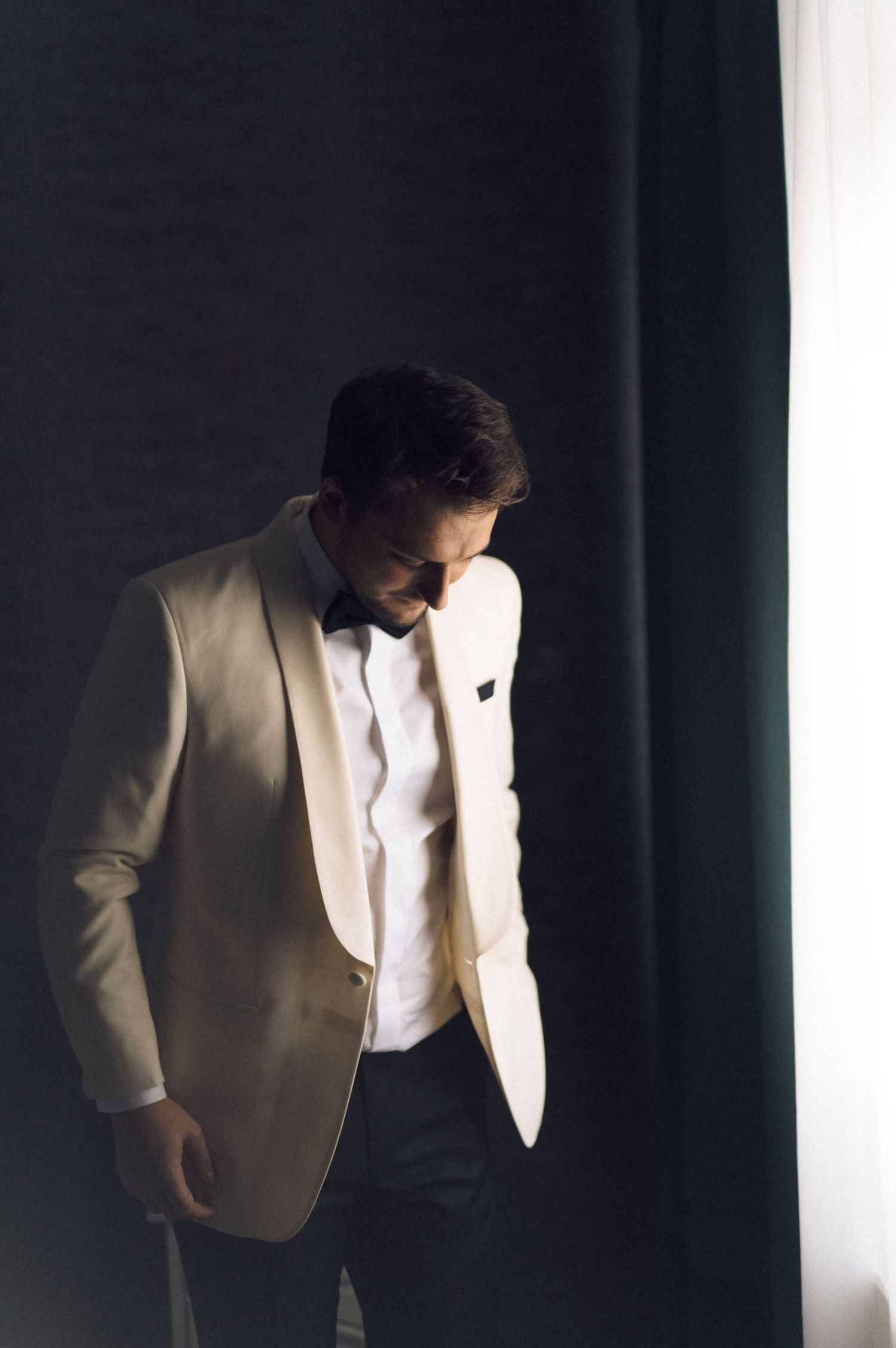 A person in a cream tuxedo jacket and black bow tie stands in dramatic lighting against a dark background.