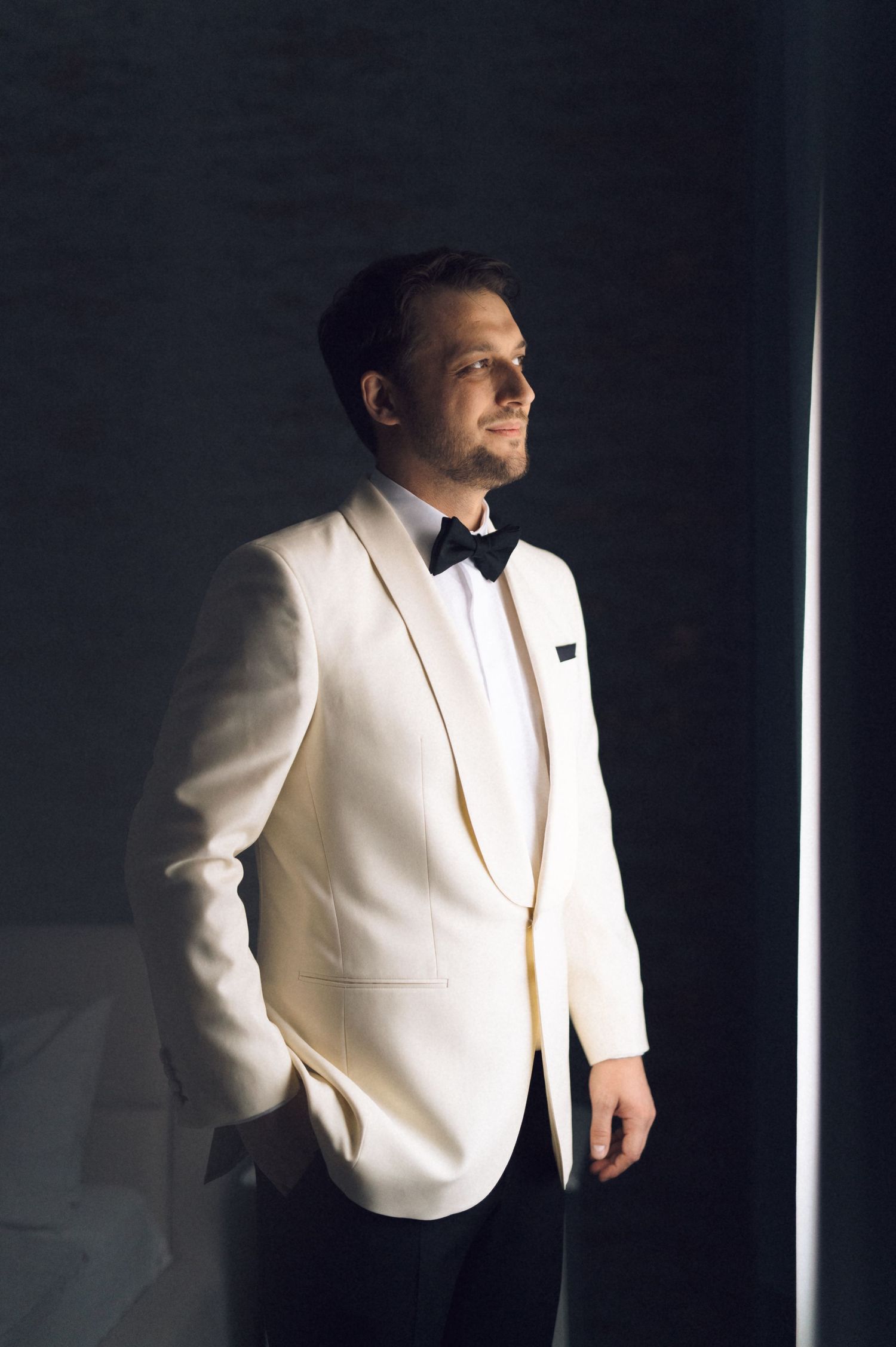 A person wearing a white tuxedo jacket and bow tie stands in dramatic lighting near a window.