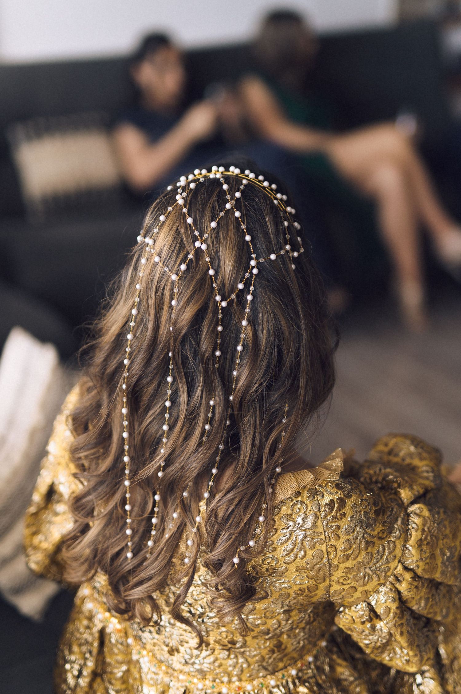 Intricate pearl hair accessories woven through wavy brown hair, with person wearing a gold textured dress in background.
