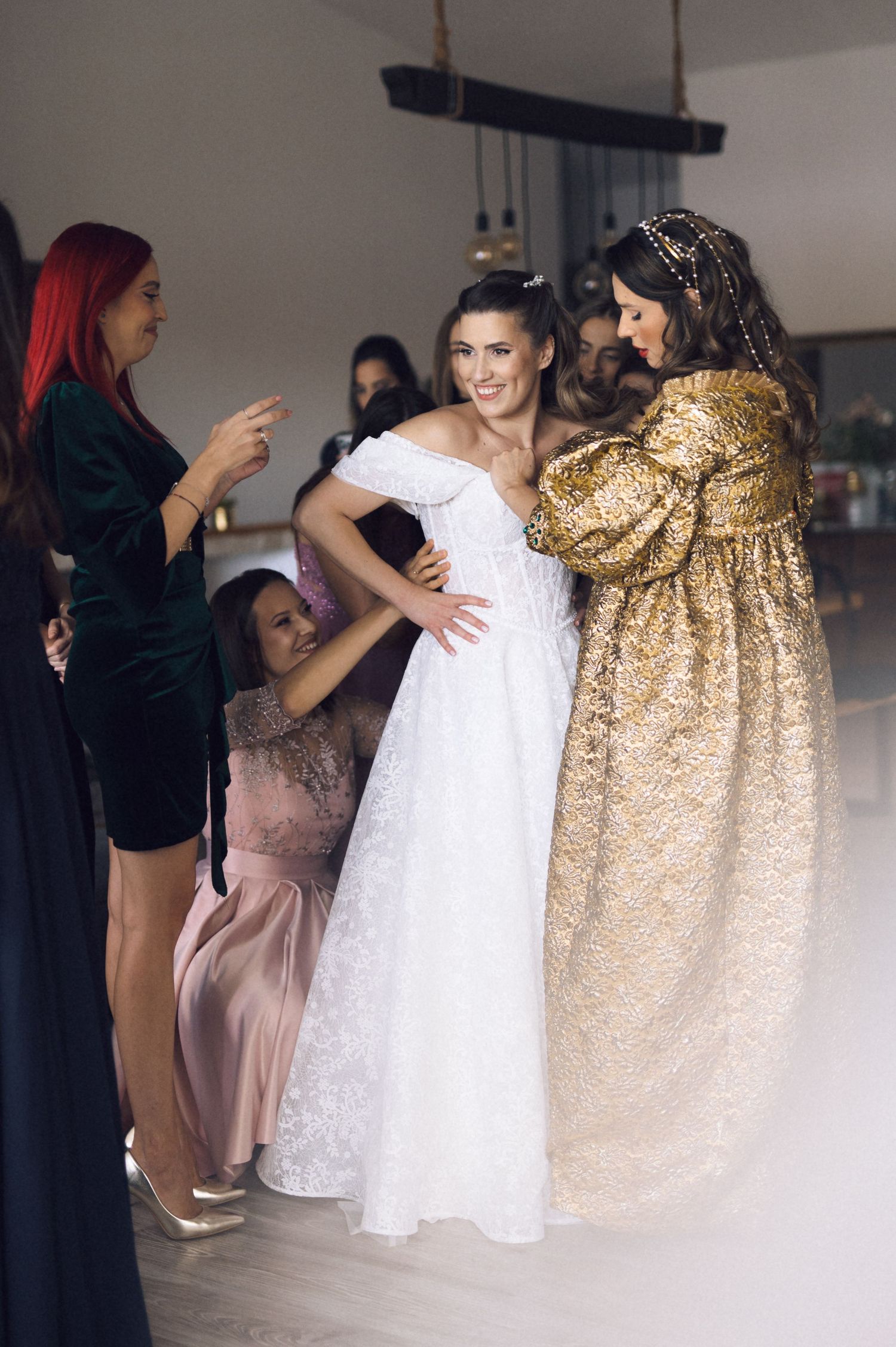 A group of people help adjust a white wedding dress during a fitting session, with someone wearing a sparkly gold outfit nearby.