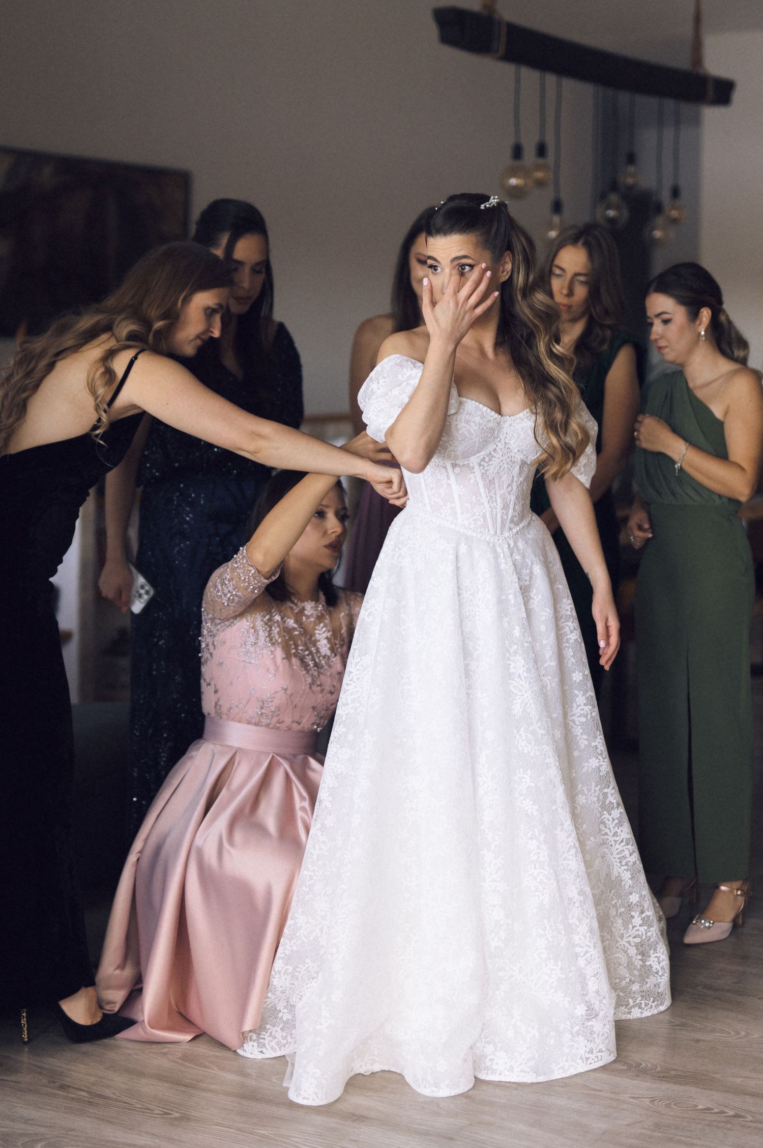 A bride in a white lace gown shares an emotional moment with bridesmaids in pink and dark formal dresses.
