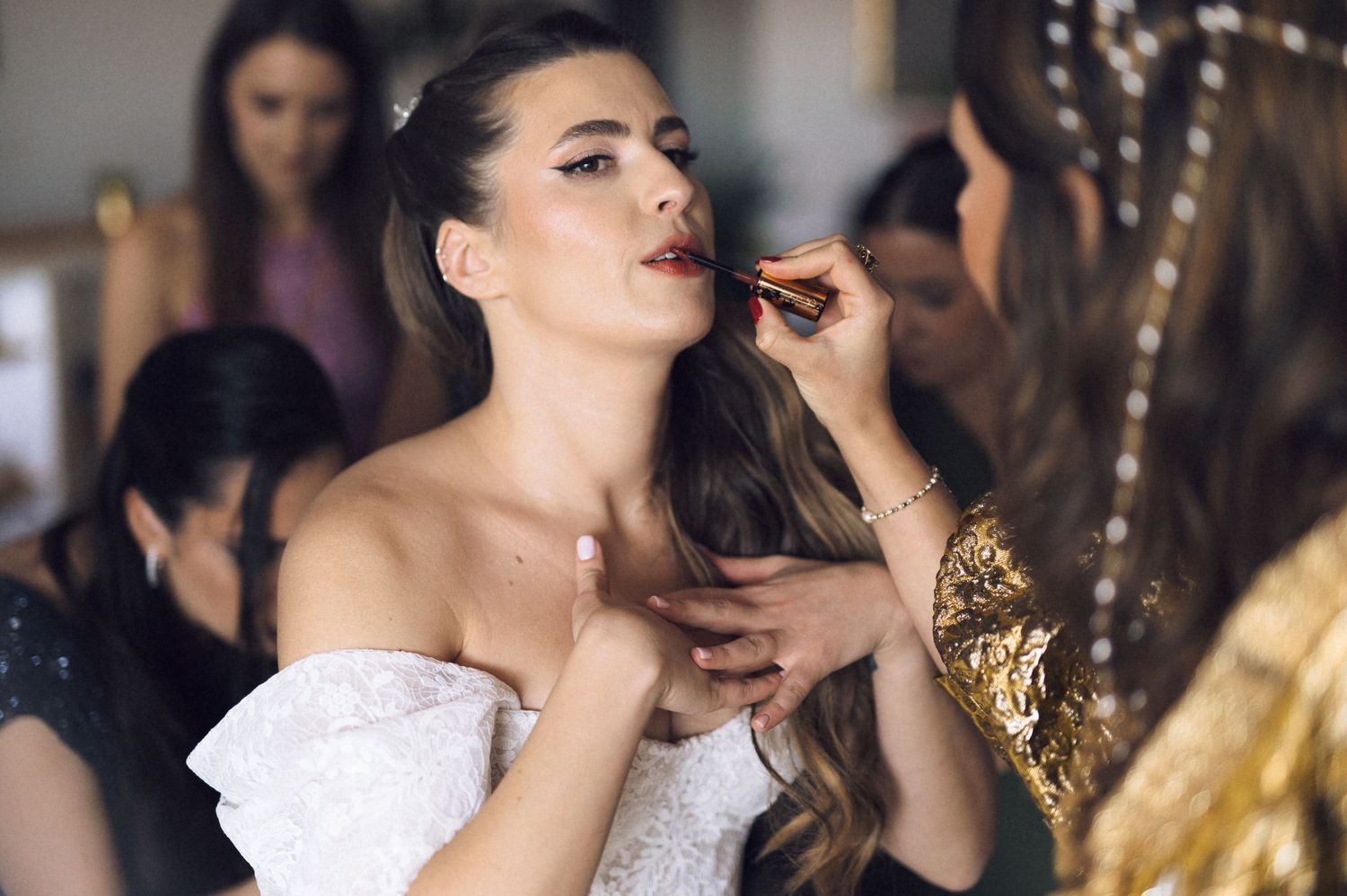 A bride in a white strapless dress gets her makeup applied during wedding preparations.