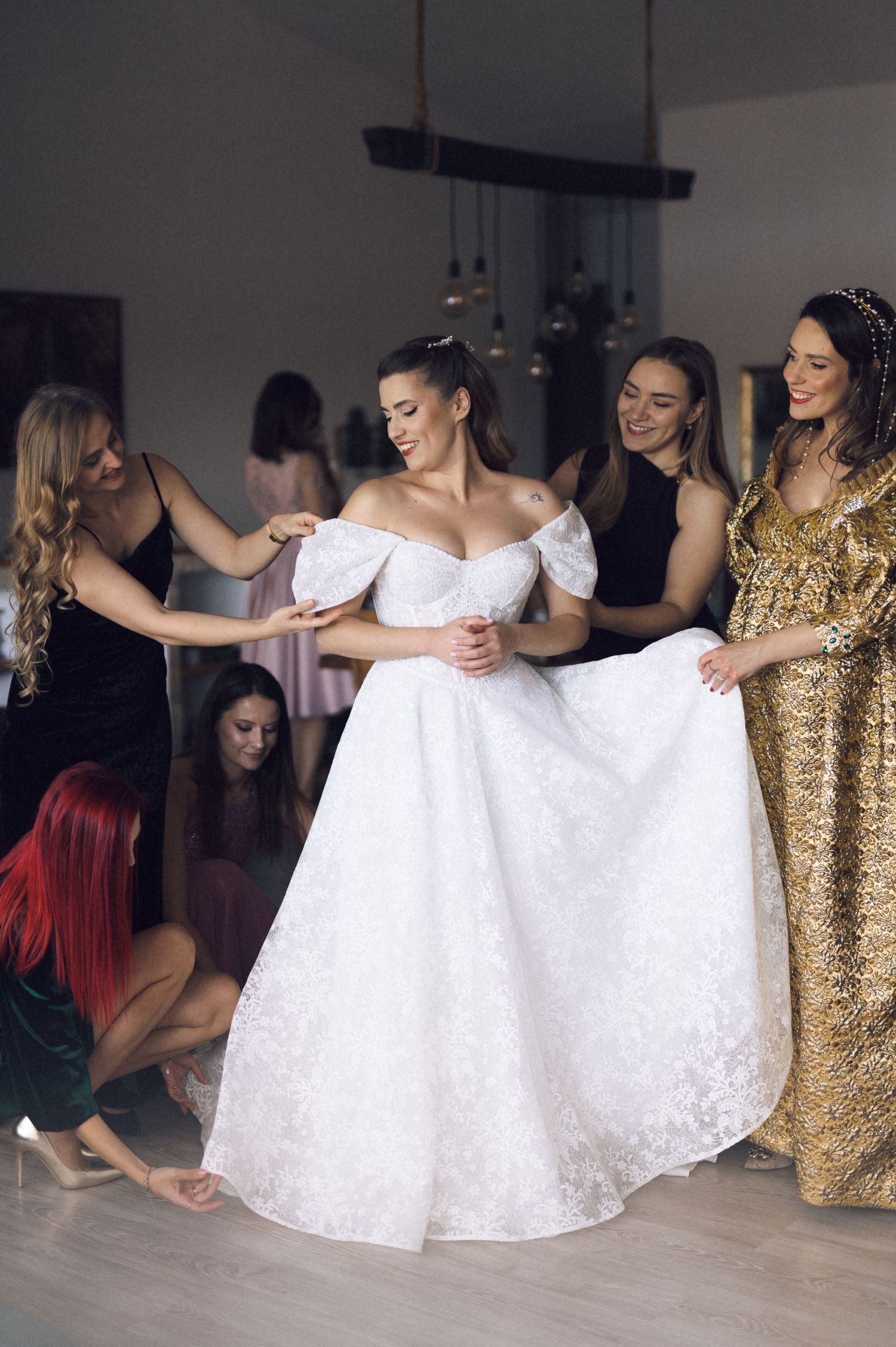 A bride in a white off-shoulder wedding gown is helped by friends in formal dresses during wedding preparations.