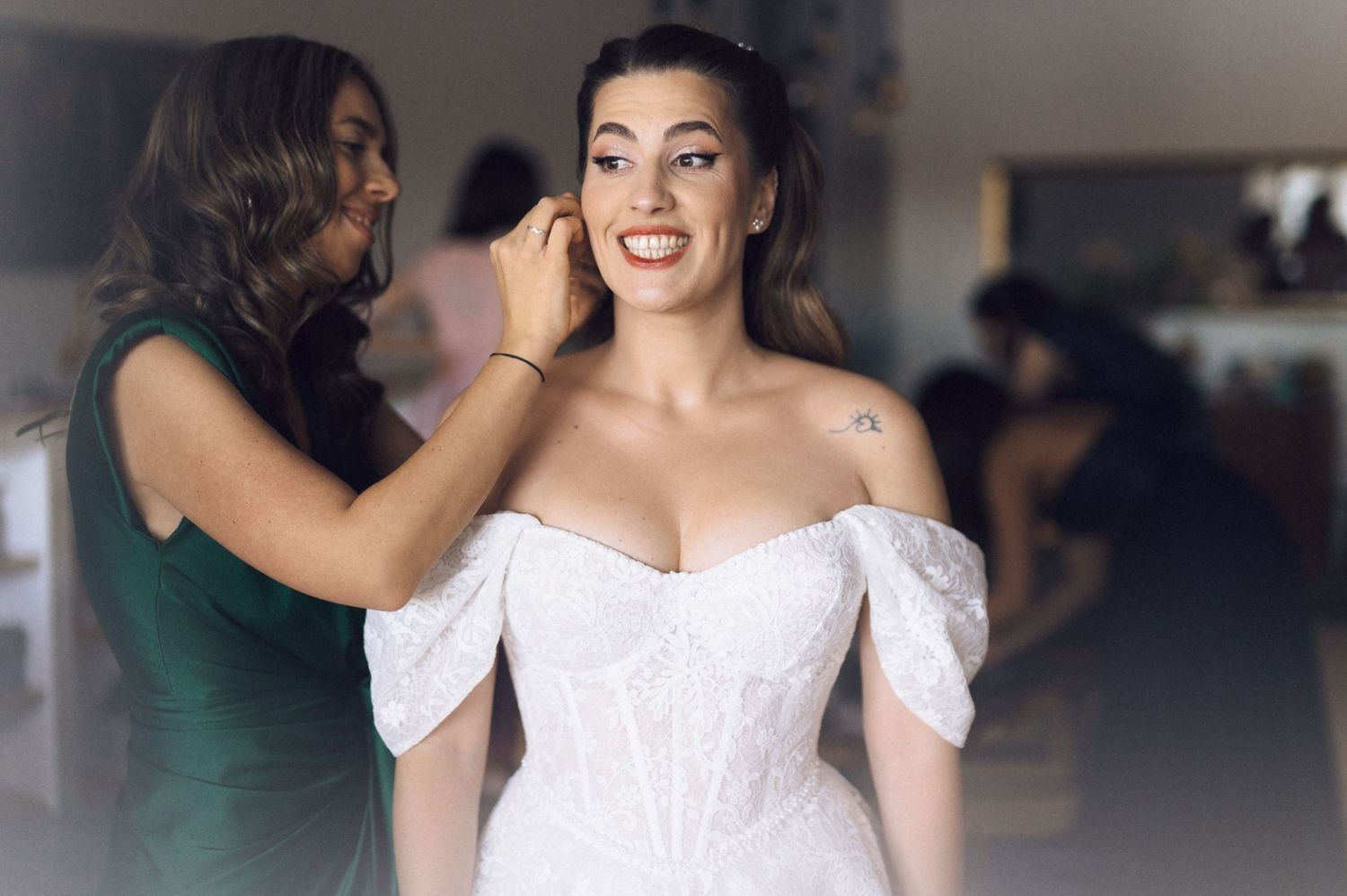 Makeup artist applies finishing touches to a smiling person wearing an elegant white off-shoulder wedding dress.
