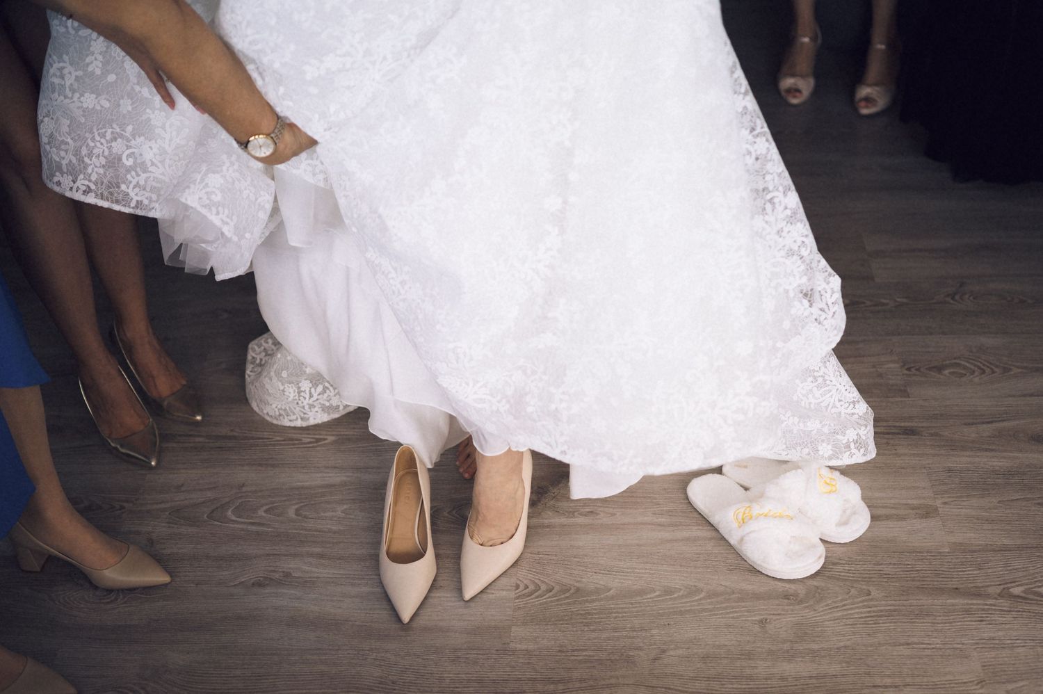Close-up view of elegant bridal shoes and white wedding dress hems on a wooden floor.