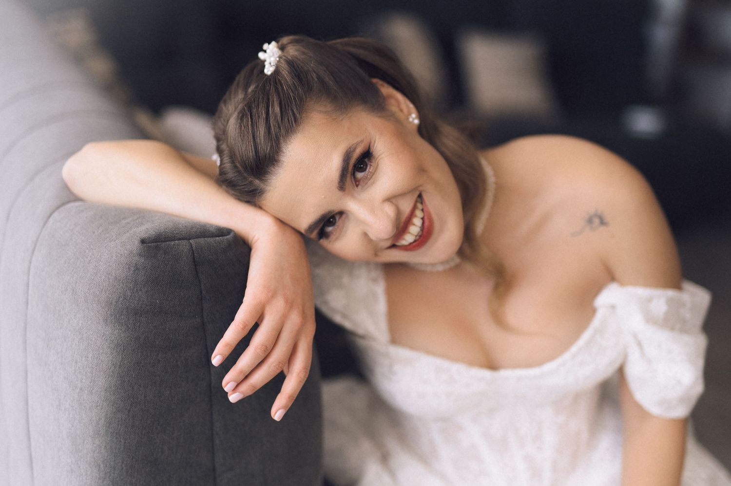 A bride in a white off-shoulder wedding dress poses elegantly in a romantic indoor setting.