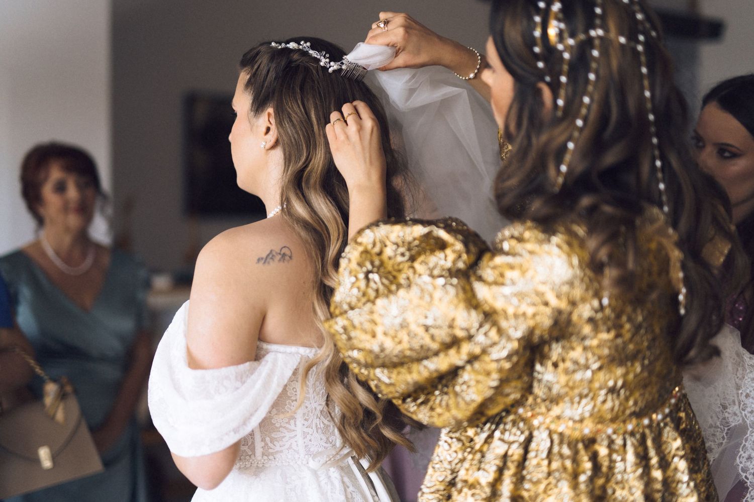 A bride in an off-shoulder white dress gets her hair styled while someone in a gold sequined dress stands nearby.