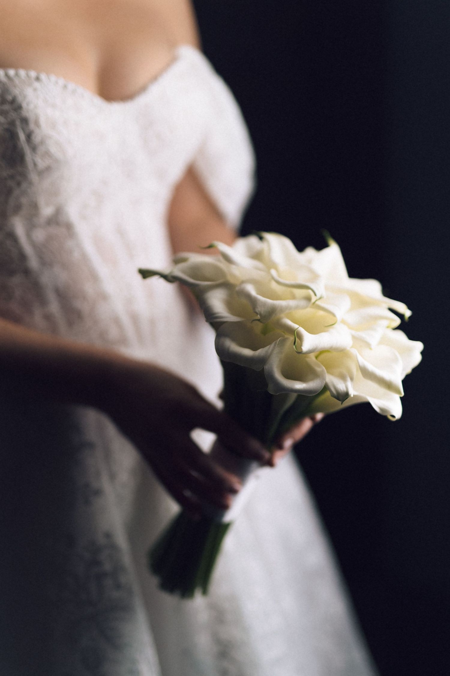 A white calla lily bridal bouquet held against a white wedding dress in dramatic lighting.