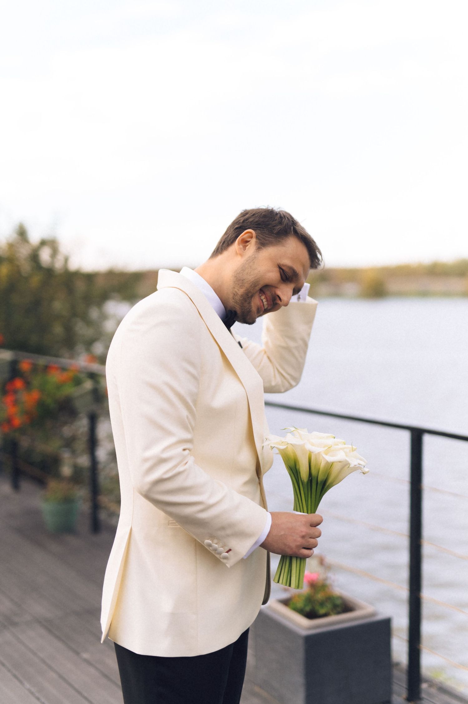 A person in a cream colored suit jacket holds a white floral bouquet against a waterfront backdrop.