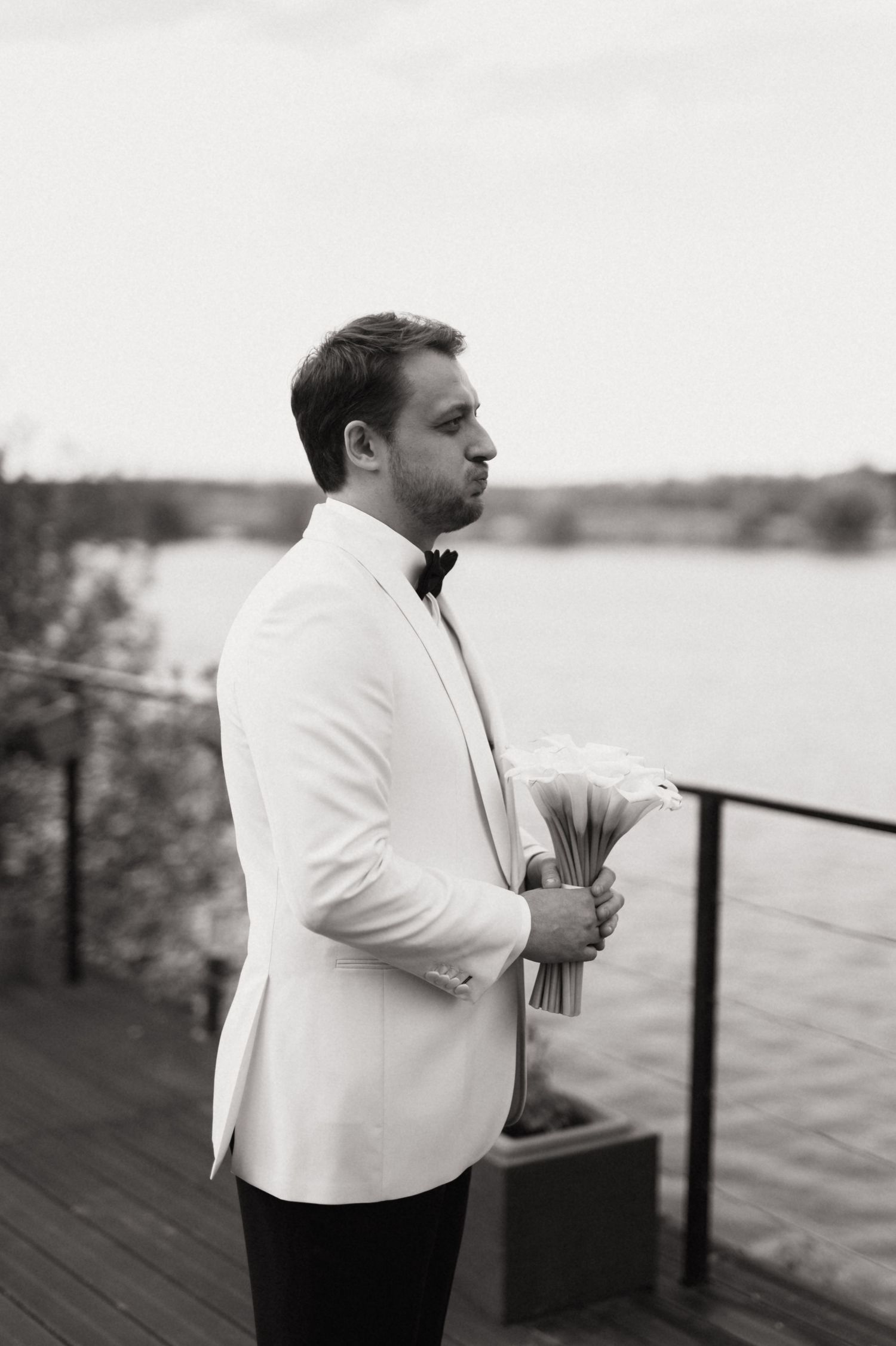 A groom in a white dinner jacket and bow tie holds a bouquet while standing on a waterfront deck.