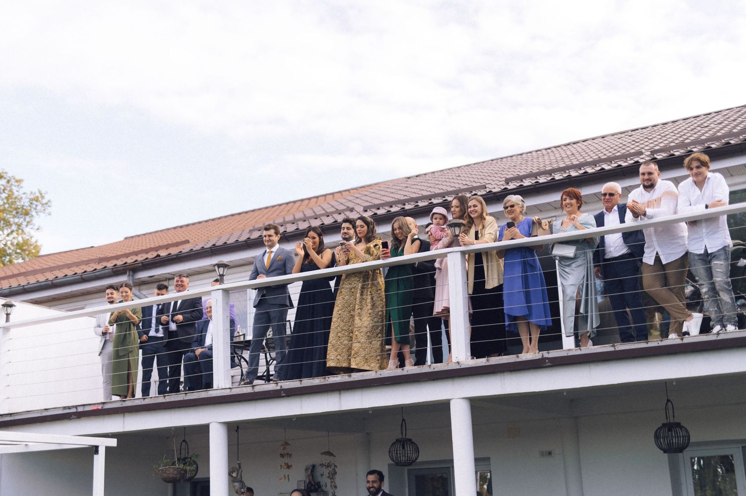 A large group of people stand along a white balcony railing overlooking an outdoor area below.