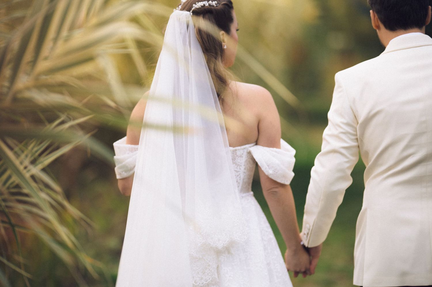 A bride and groom hold hands while walking through a scenic outdoor setting with palm fronds in the background.