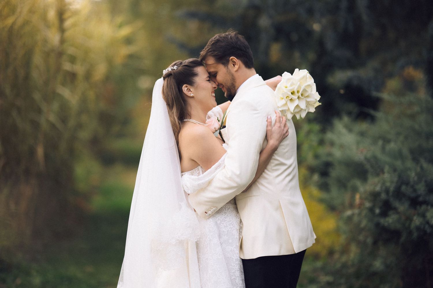 A romantic wedding couple embraces outdoors with the bride holding a white bouquet against a dreamy natural backdrop.