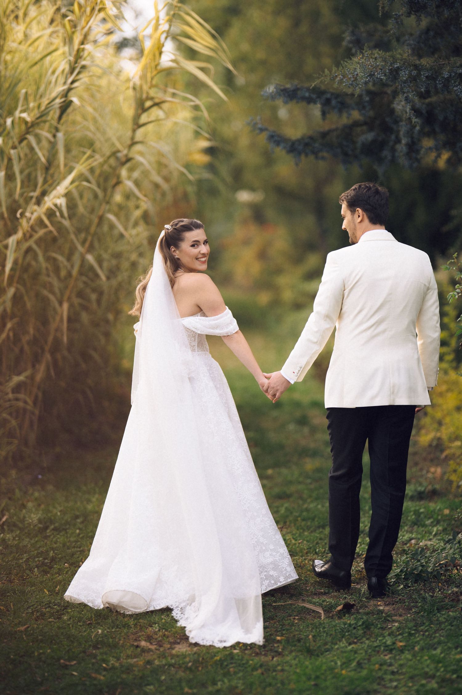 A couple in wedding attire walks hand in hand through a garden path with tall grass in the background.