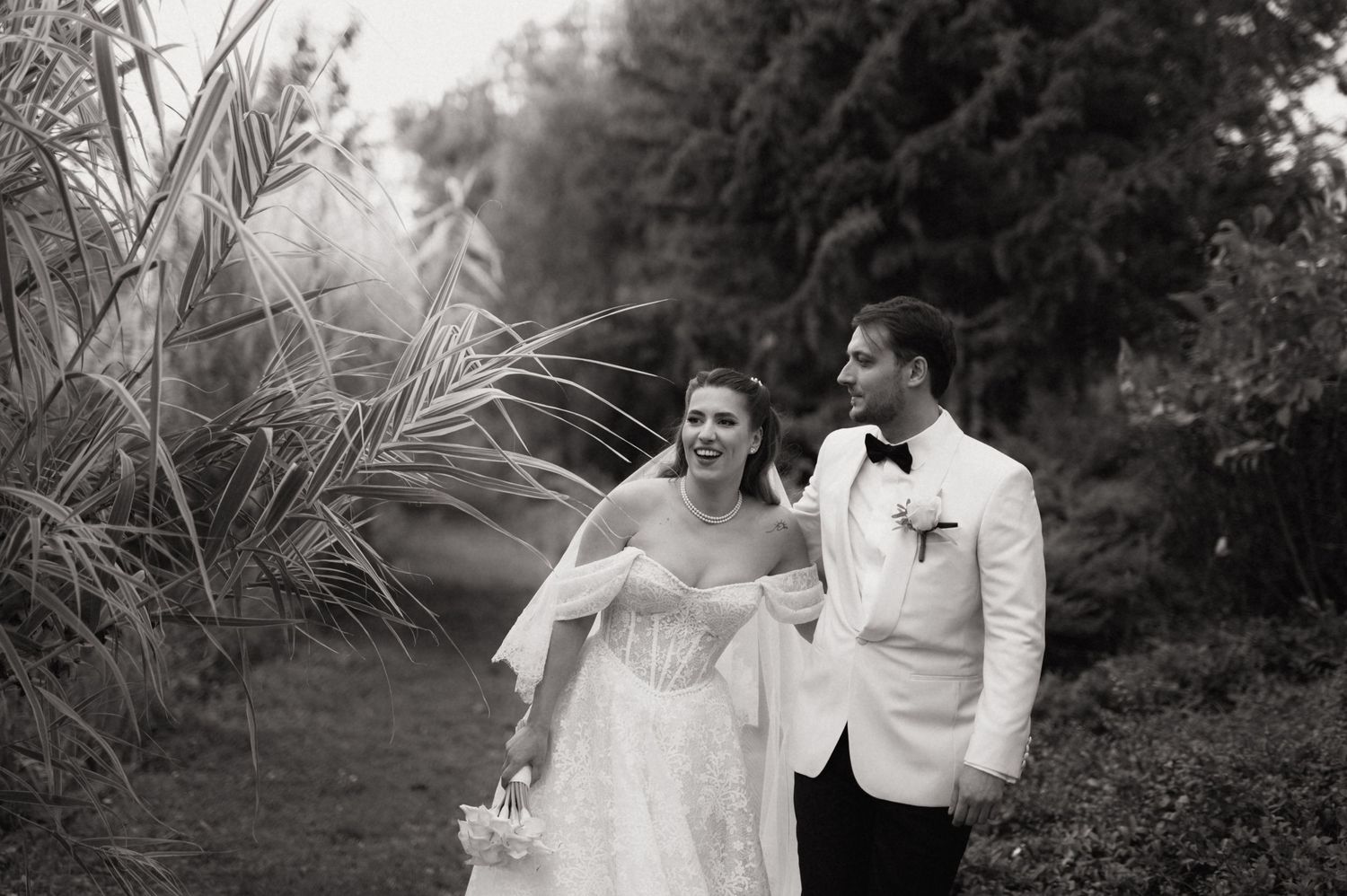 A couple in wedding attire walks together outdoors along a garden path in black and white photography.