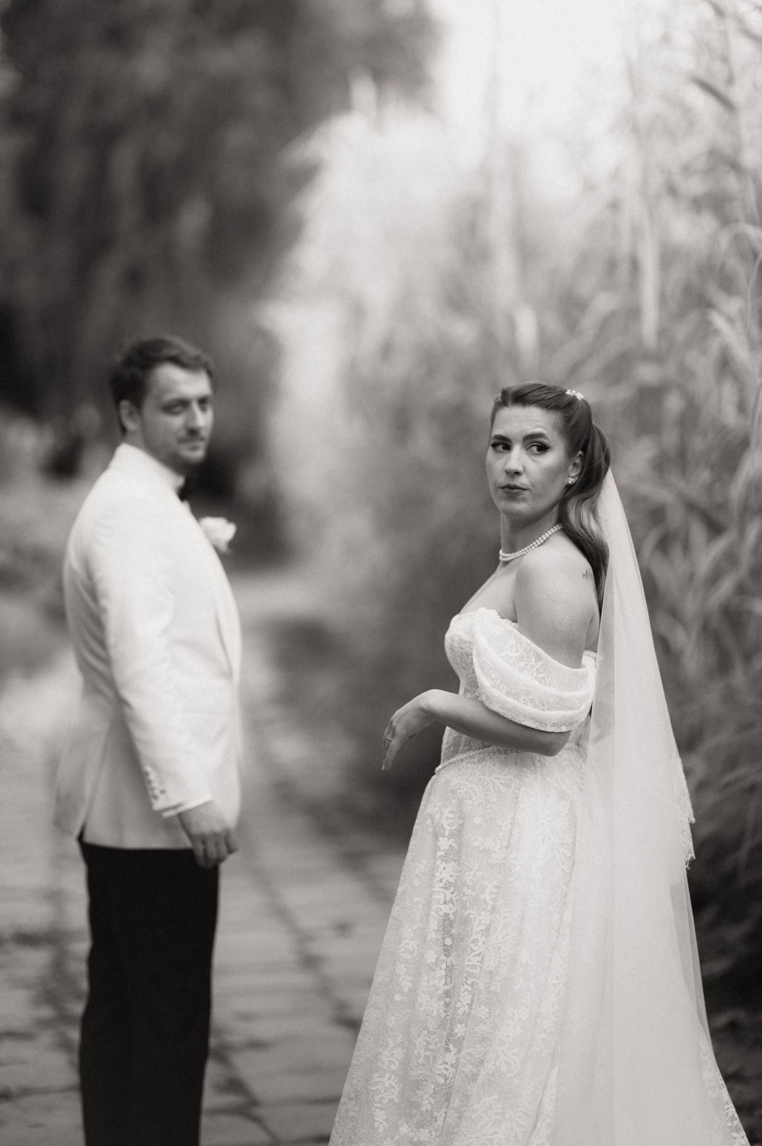 A black and white wedding photo showing a bride in a flowing white dress and veil with groom in white jacket on garden path.