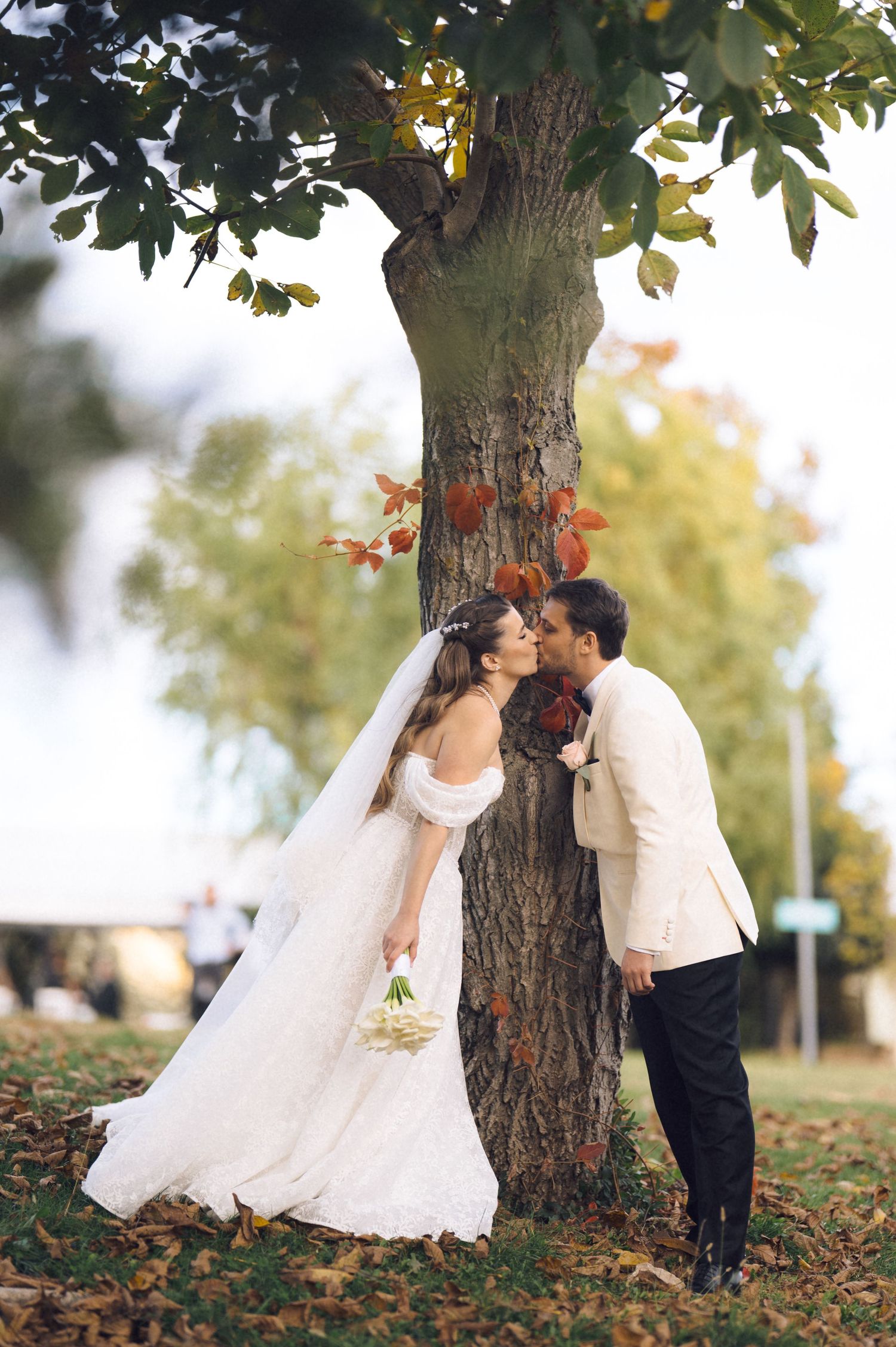 Romantic wedding couple shares tender moments under autumn trees with fallen leaves in a park setting.