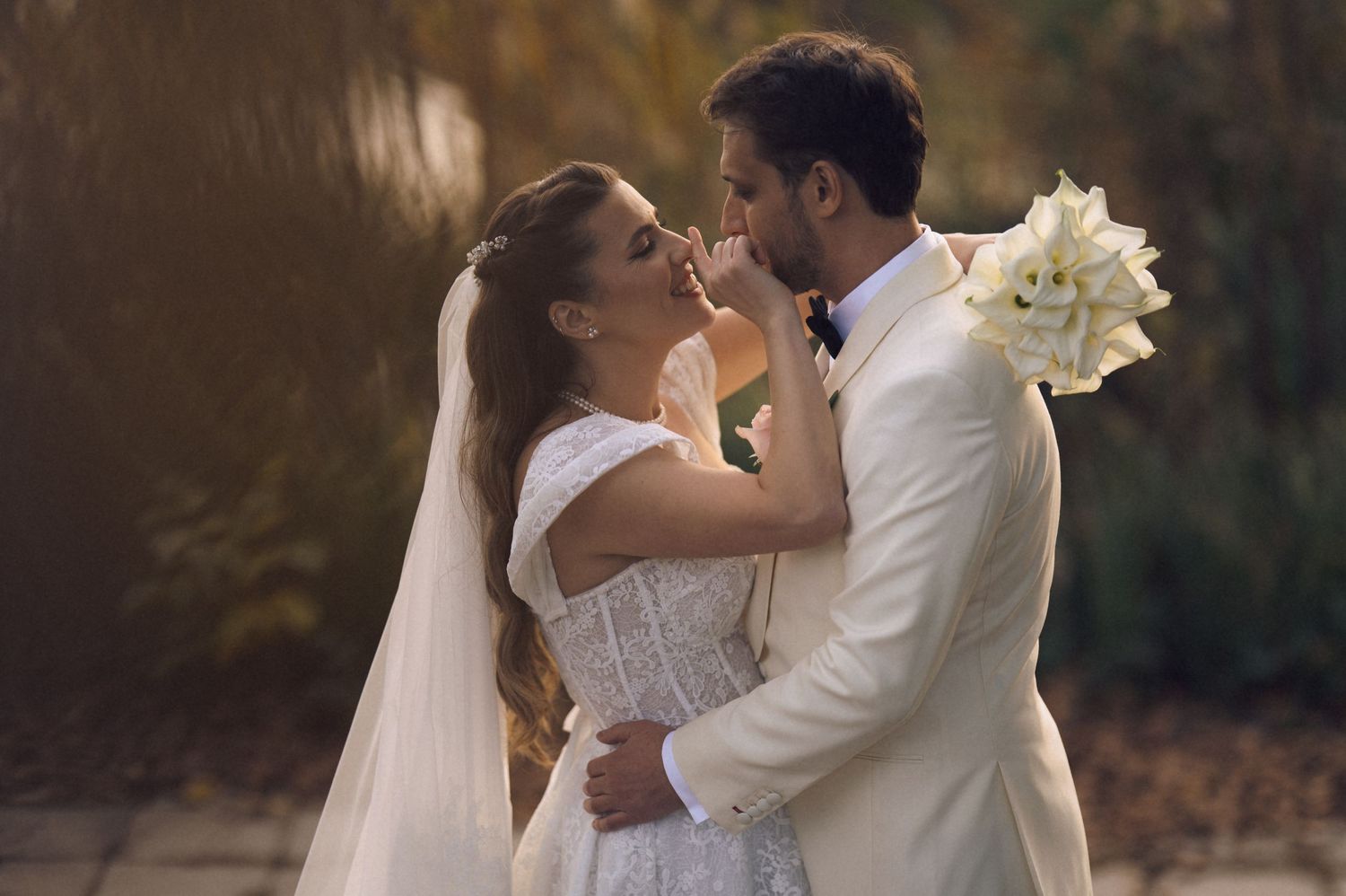 A romantic wedding couple embraces outdoors at sunset while the bride holds a bouquet of white flowers.