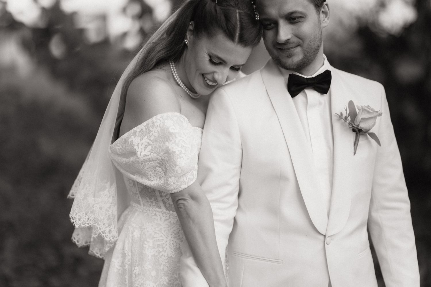 A romantic black and white wedding photo showing a couple in white formal attire sharing a tender moment outdoors.