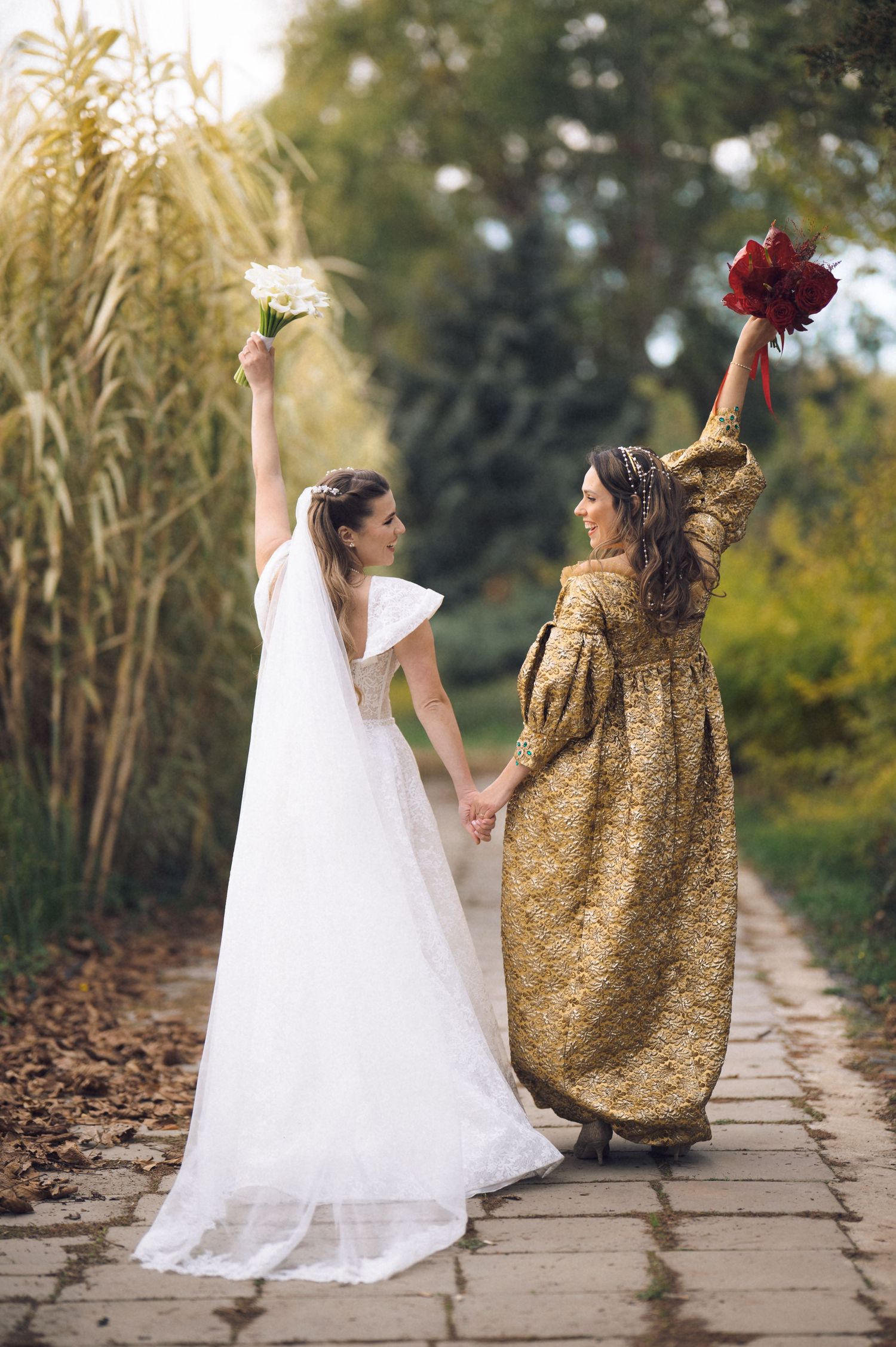 Two people in contrasting white and gold outfits dance joyfully on a stone path surrounded by autumn foliage.