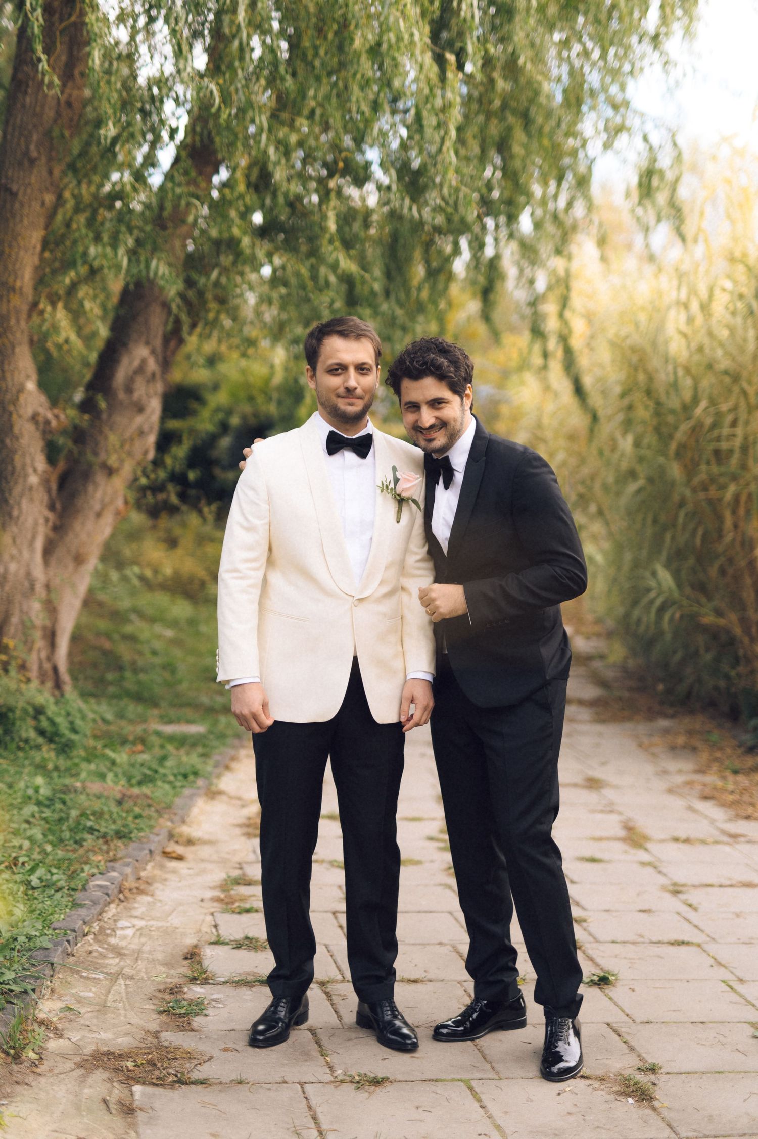 Two people in formal attire, one in white and one in black, stand together on a stone path surrounded by trees.
