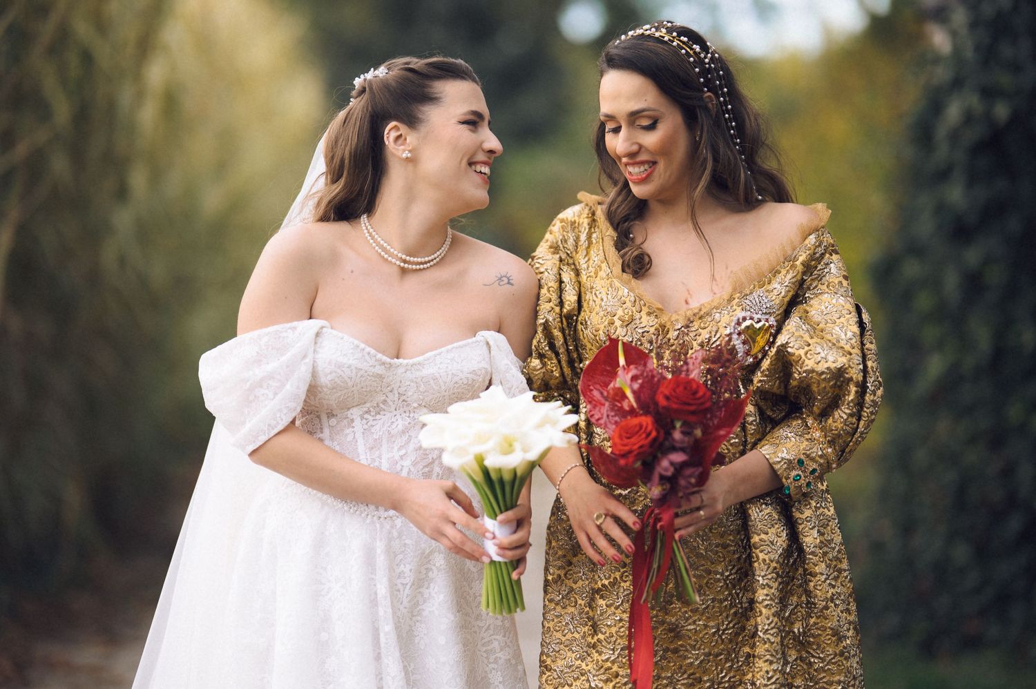 Two brides share a joyful moment outdoors, one wearing white and holding white flowers, the other in gold with red flowers.